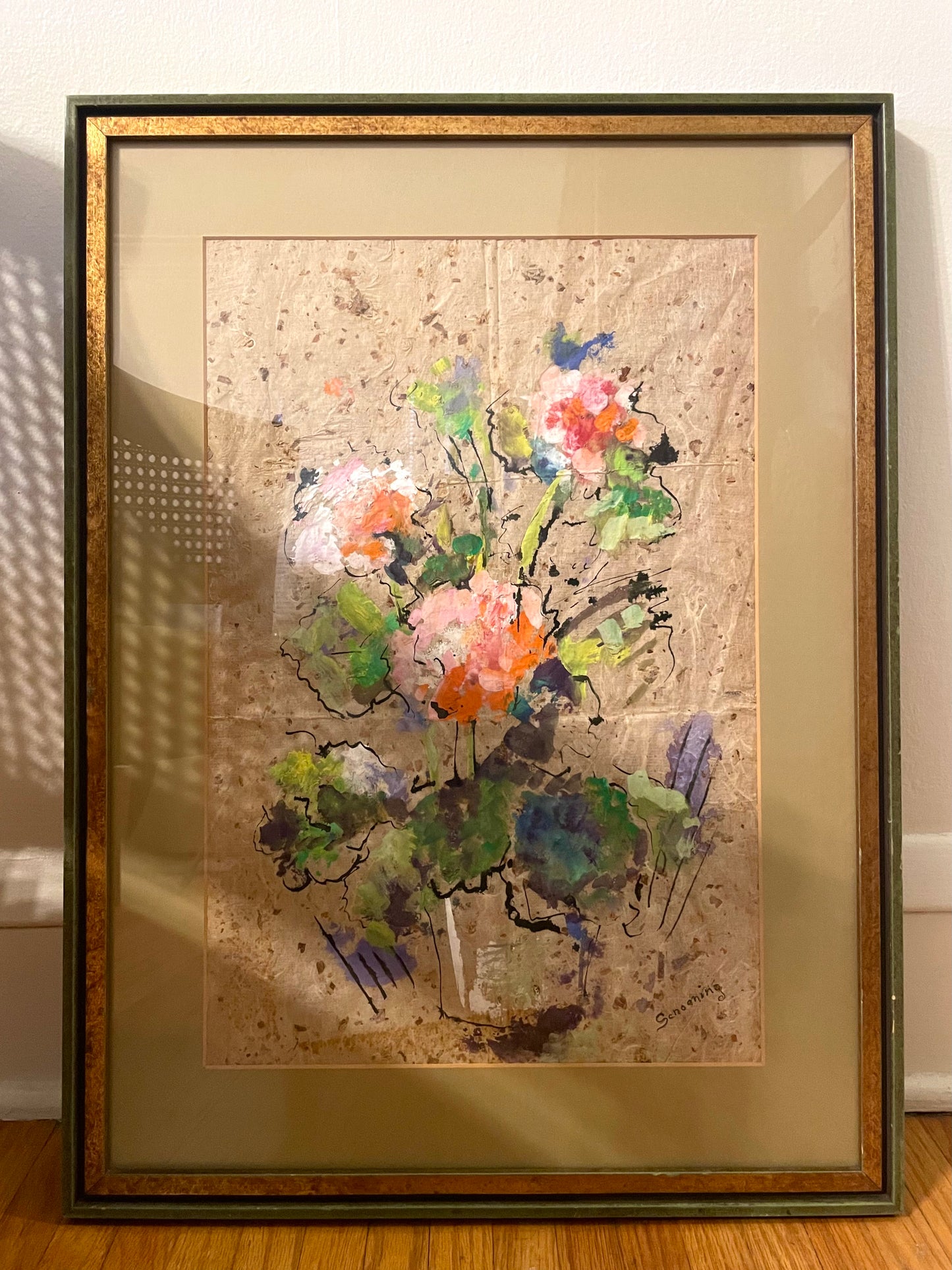 Framed expressionist painting of a floral bouquet on textured beige paper, set in a gold-toned and green frame with a beige mat, leaning against a white wall on a wooden floor.