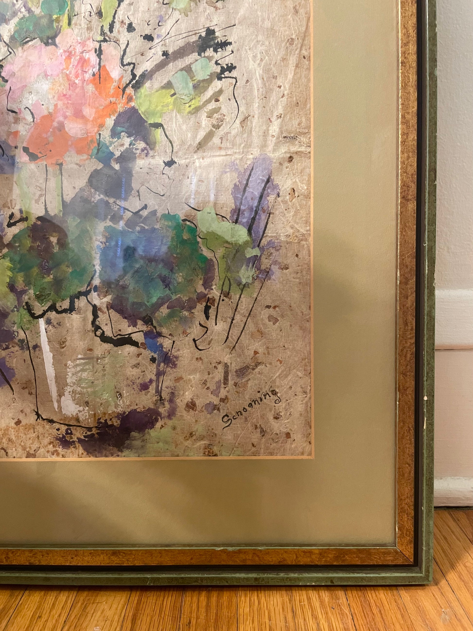 A framed abstract painting with a textured beige background and expressive brushstrokes in green, blue, pink, orange, and purple, shown resting on a wooden floor against a white wall.
