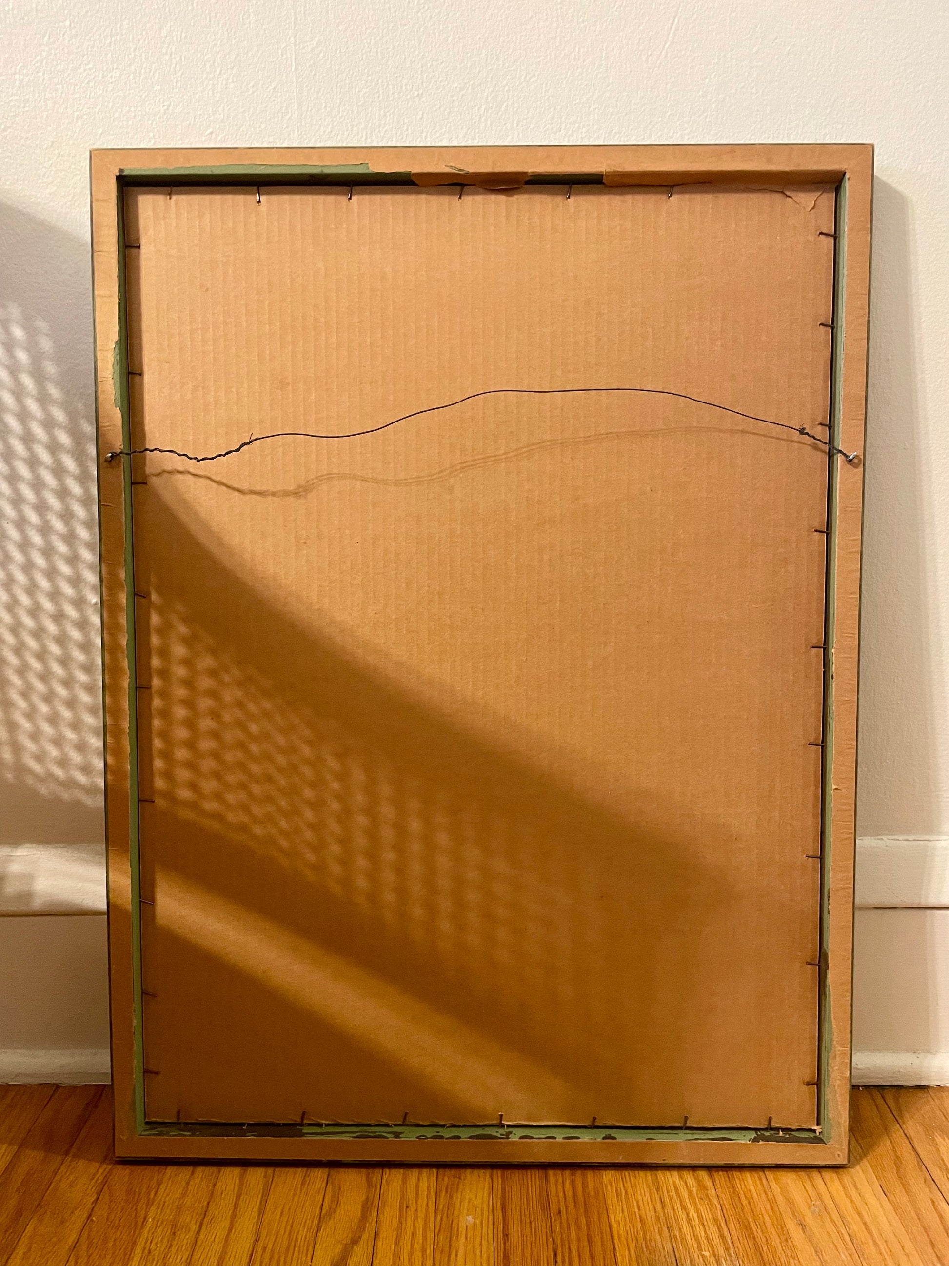 The back side of a framed artwork leaning against a white wall on a wooden floor. The frame is light brown wood, and the backing is tan cardboard with visible hanging wire.