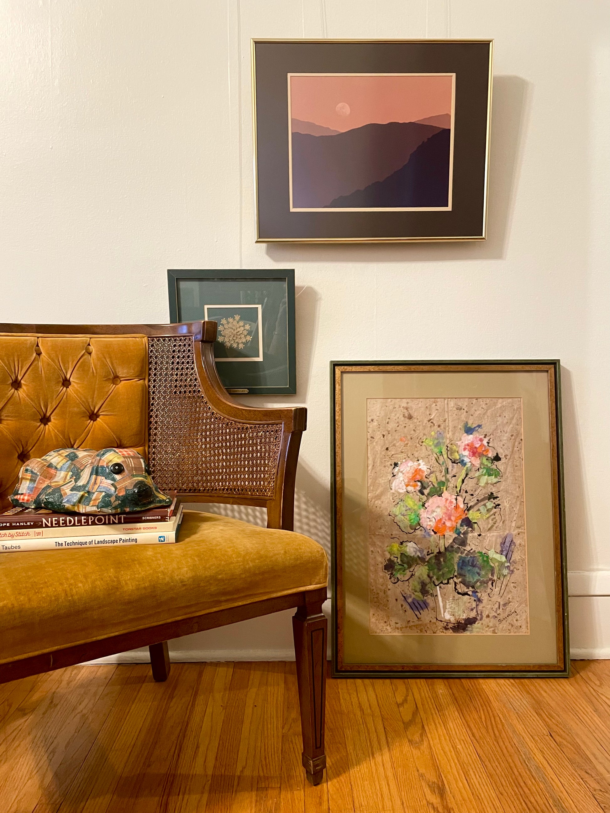 A styled interior scene showing framed wall art in a home setting. A mustard yellow tufted chair with a stack of books and a patchwork dog figurine sits on a hardwood floor, next to a framed floral painting leaning against the wall, with a framed landscape photograph depicting the silhouette of a mountain range under a pink and orange sky with a moon in the sky hanging on the wall.