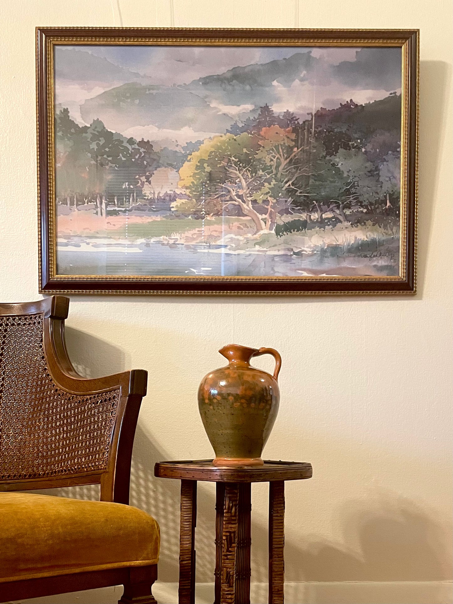 A framed landscape watercolor print hangs on a beige wall above a small wooden side table holding a green and orange ceramic pitcher, with part of a wooden armchair visible at the left edge.