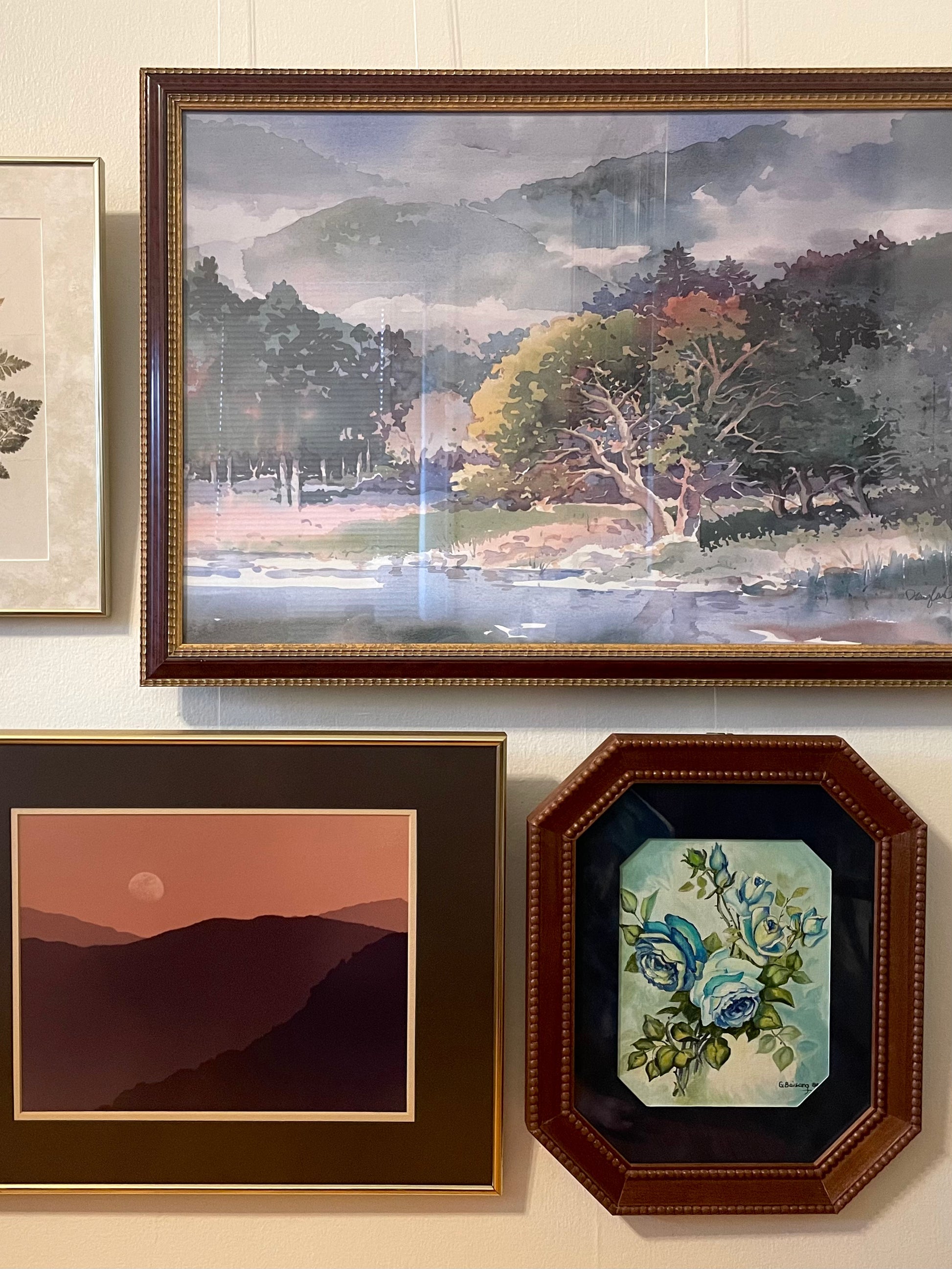 Three framed wall art pieces displayed on a light beige wall. The largest piece is a landscape painting with misty mountains, trees, and a river, in a dark brown frame.