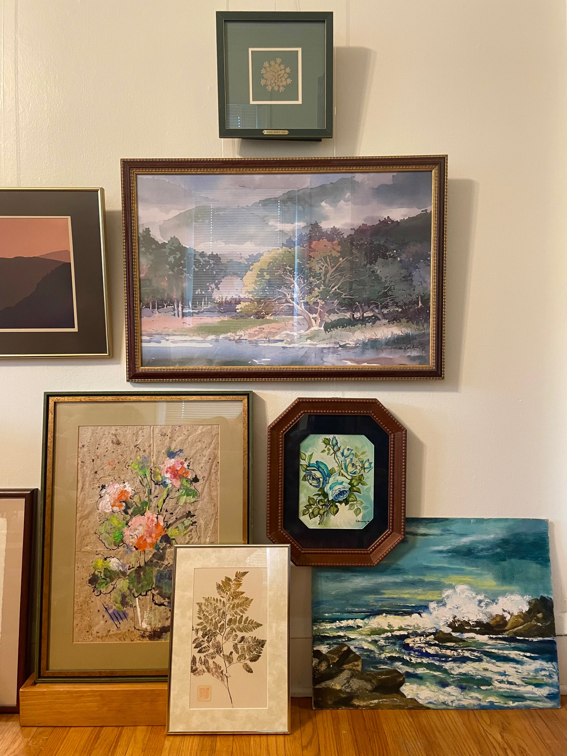 A collection of framed and unframed art pieces displayed on a beige wall above a wooden floor, including a landscape, an octagonal floral painting, an ocean scene, and several pressed and framed botanicals.