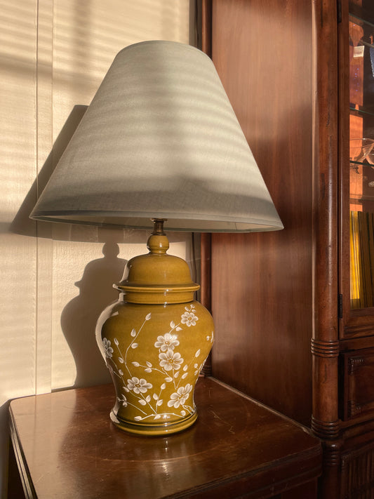 A table lamp with a tapered light blue fabric shade and a glossy olive green ceramic base decorated with white floral patterns, set on a dark wooden side table in a warm indoor setting.