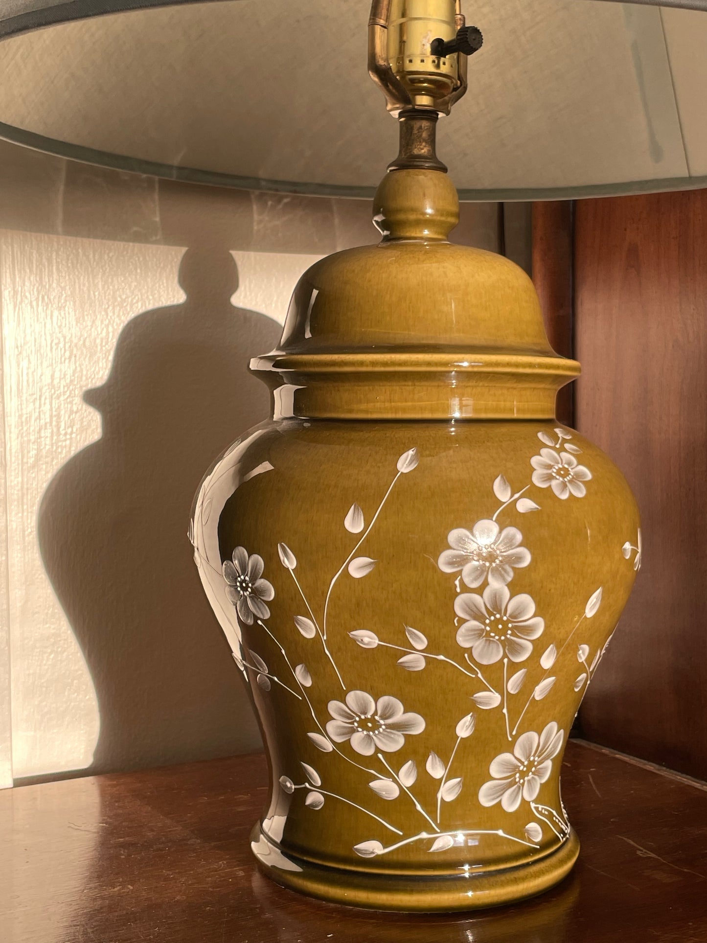 A table lamp with a tapered light blue fabric shade and a glossy olive green ceramic base decorated with white floral patterns, set on a dark wooden side table in a warm indoor setting.