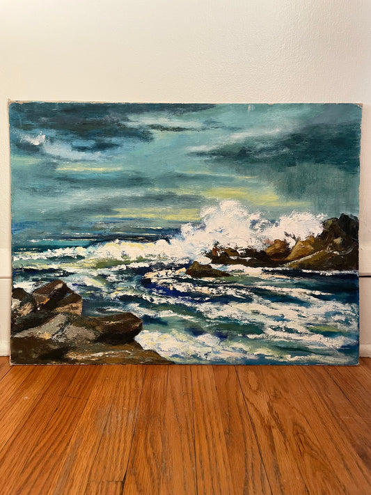 An oil painting of a rocky coastline with crashing waves under a stormy sky, displayed on a wooden floor against a white wall.