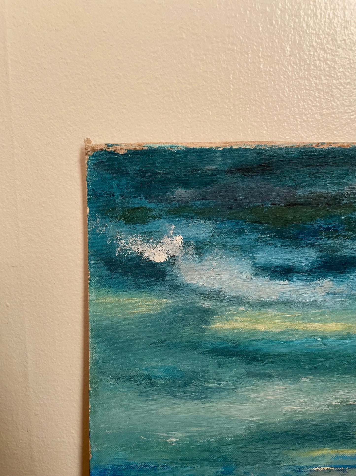 A close-up of the upper left corner of a painting depicting an ocean scene in shades of teal, blue, green, and white, leaning against a white wall. Wear is shown at the corner of the board.