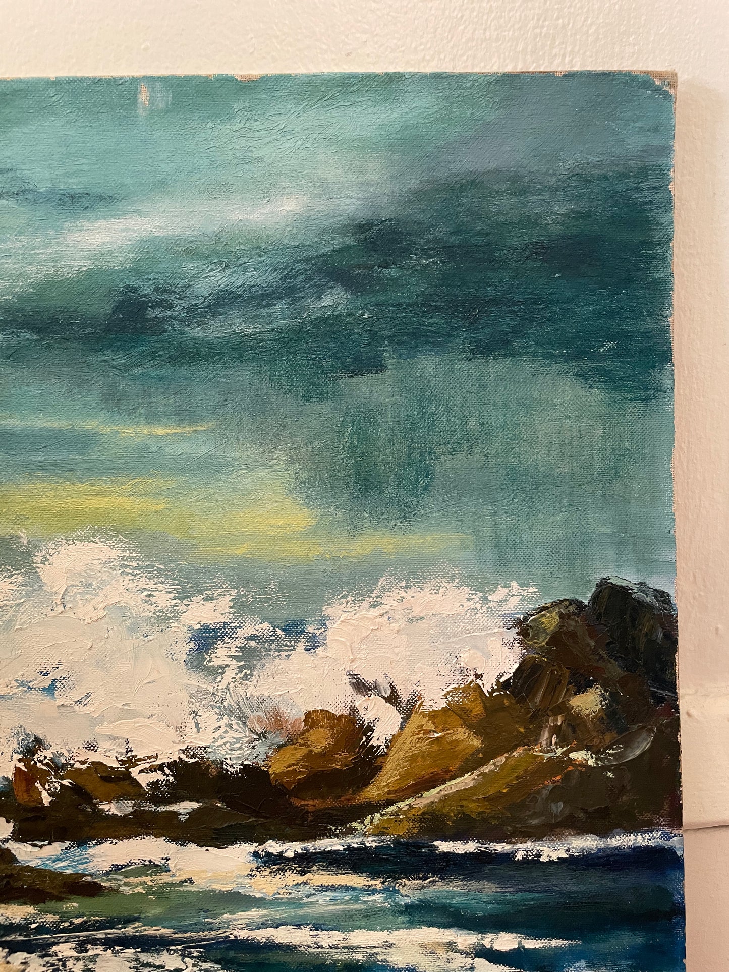 An oil painting of a dramatic coastal scene with dark teal and green stormy sky, white crashing waves, and brown rocky shoreline, shown leaning against a white wall. Wear is shown to the corner of the board.