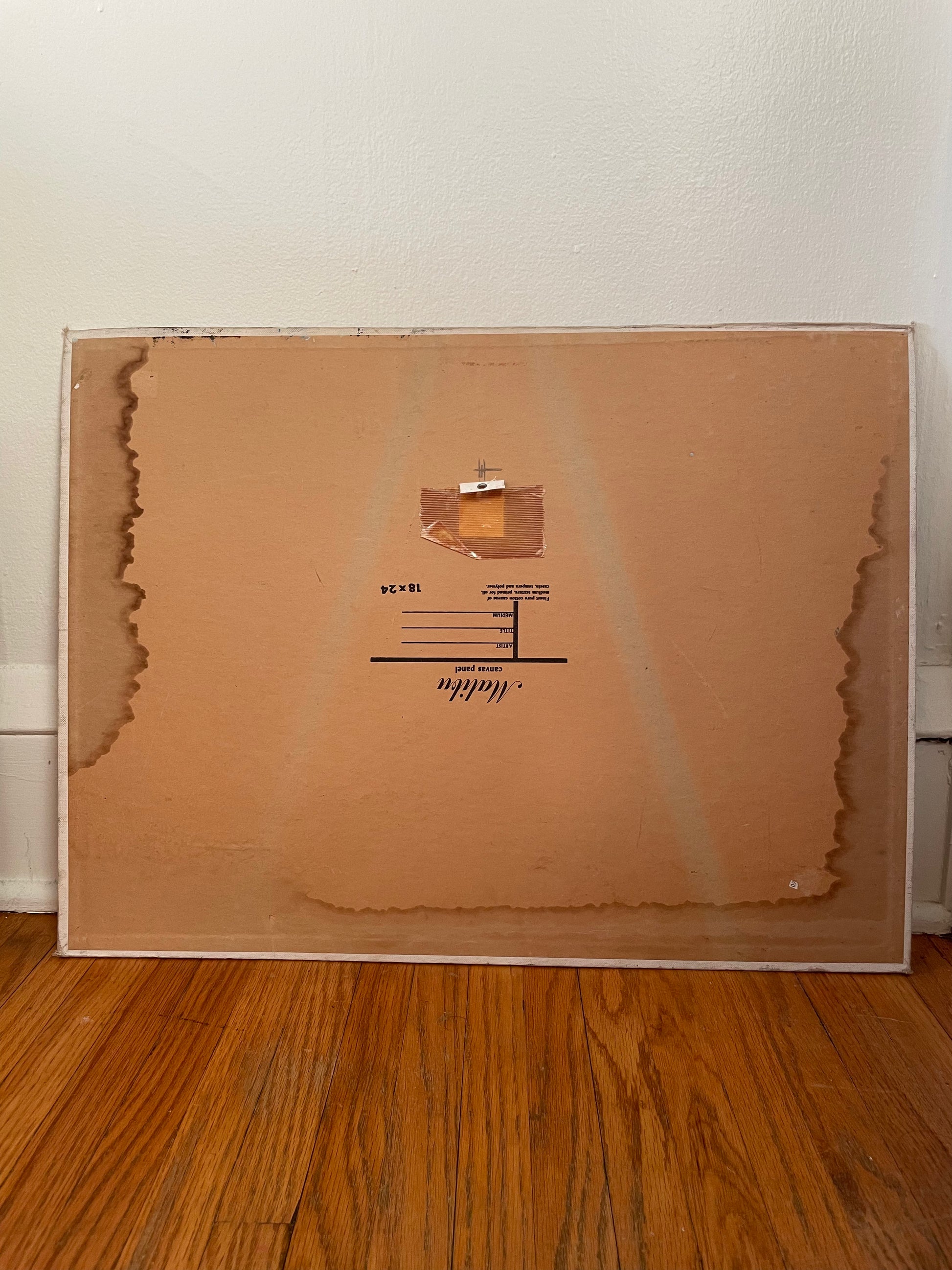 The backside of an art board is shown leaning against a white wall on a wooden floor. Some water marks are shown at the edges and there is a ring for hanging taped to the center.