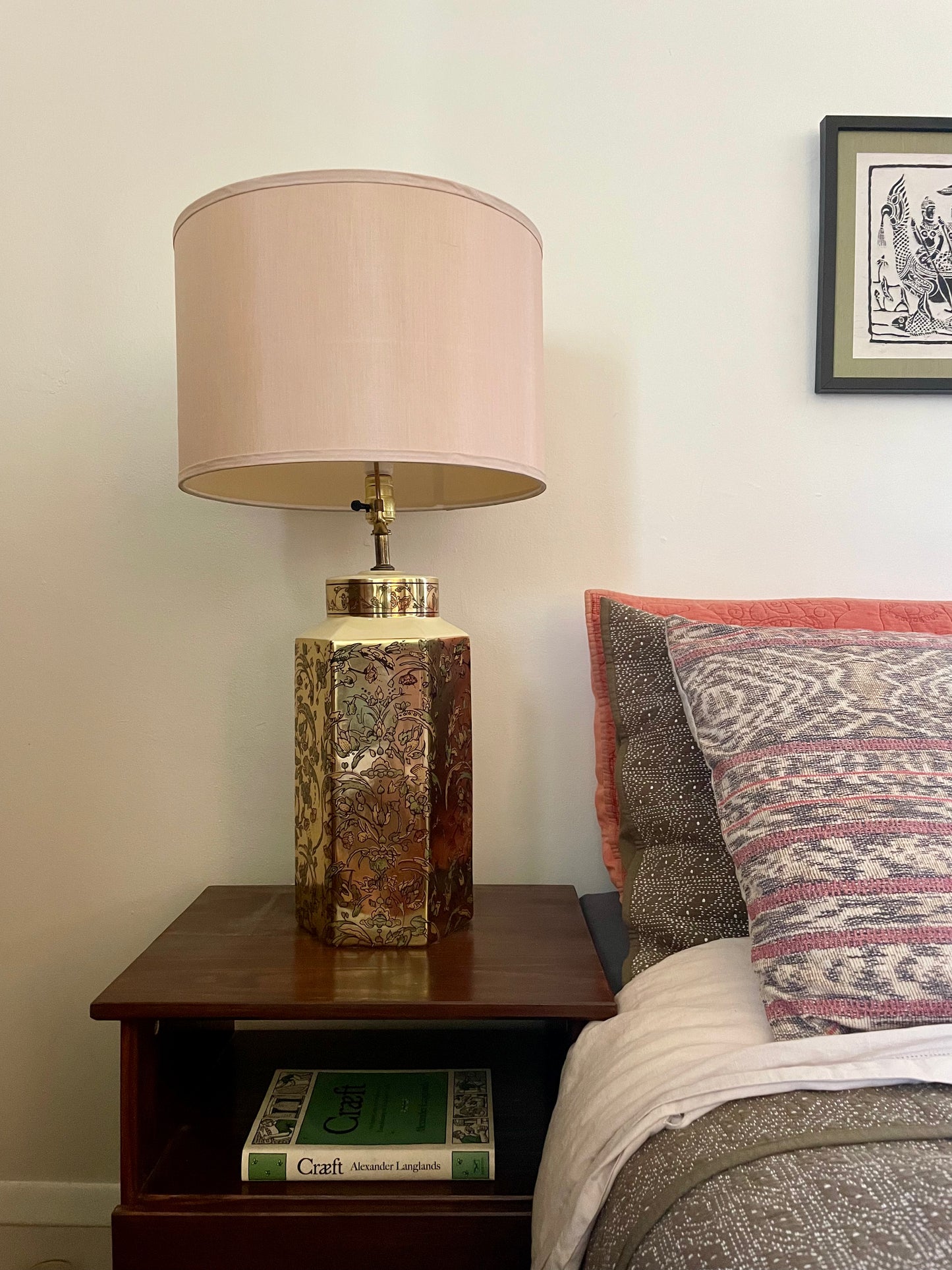 Geld hexagonal ceramic table lamp with floral pattern sitting on a dark brown nightstand next to a bed with pink and green layered pillows and a green embroidered bedspread.