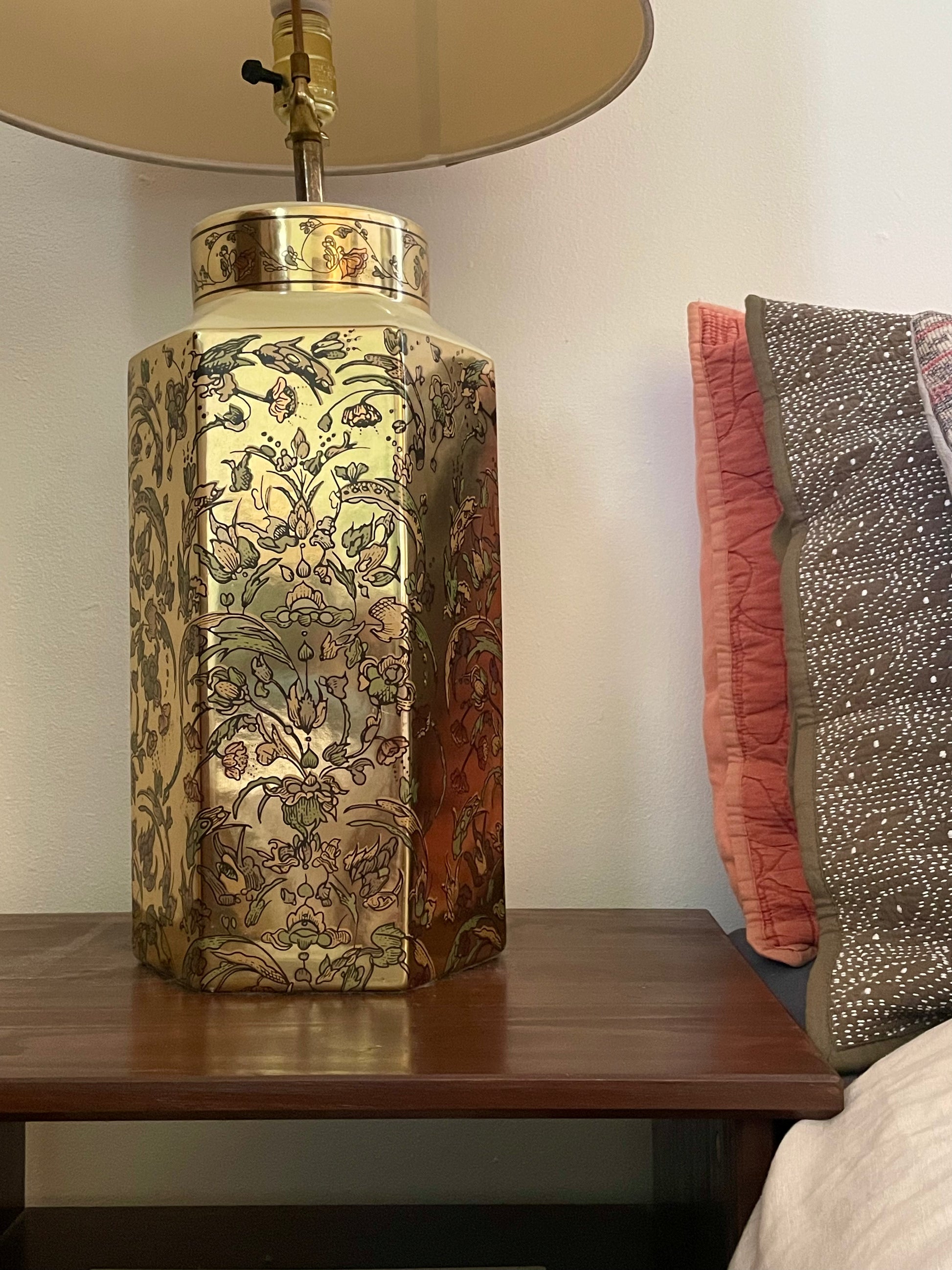 A decorative hexagonal table lamp with a gold-toned body covered in an intricate floral pattern, sitting on a dark wooden table next to a bed with patterned pillows.
