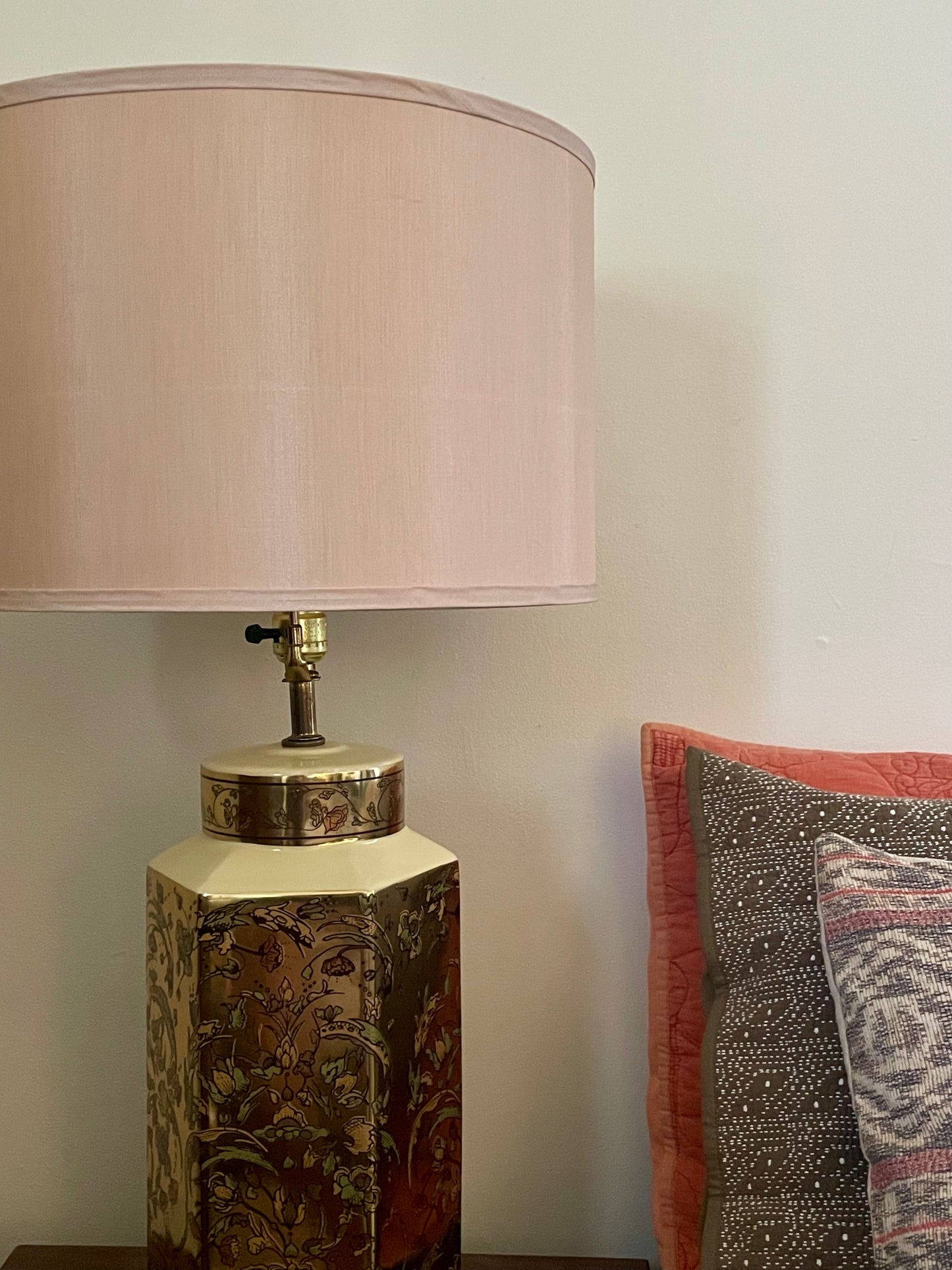 A table lamp with a taupe silk fabric drum shade and a decorative hexagonal base featuring a floral pattern in gold, green, and pink tones, shown next to patterned pillows against a plain wall.