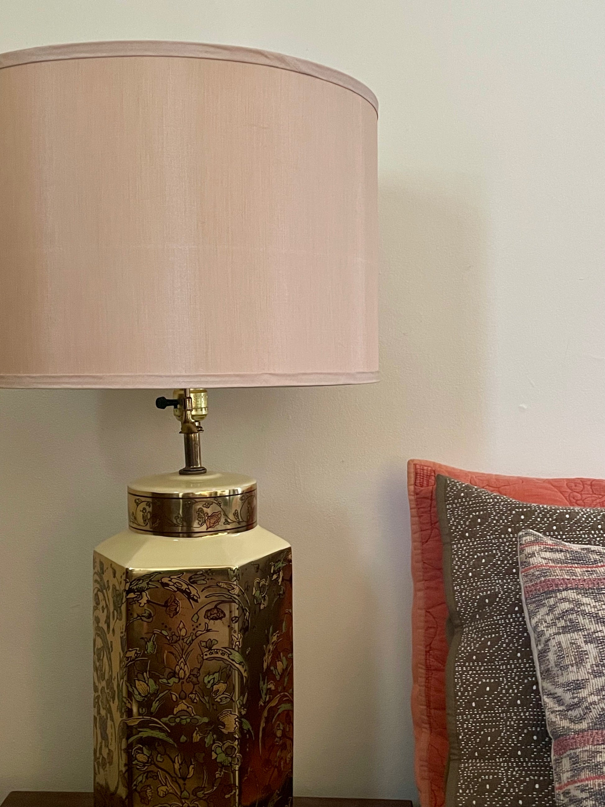 A table lamp with a taupe silk fabric drum shade and a decorative hexagonal base featuring a floral pattern in gold, green, and pink tones, shown next to patterned pillows against a plain wall.