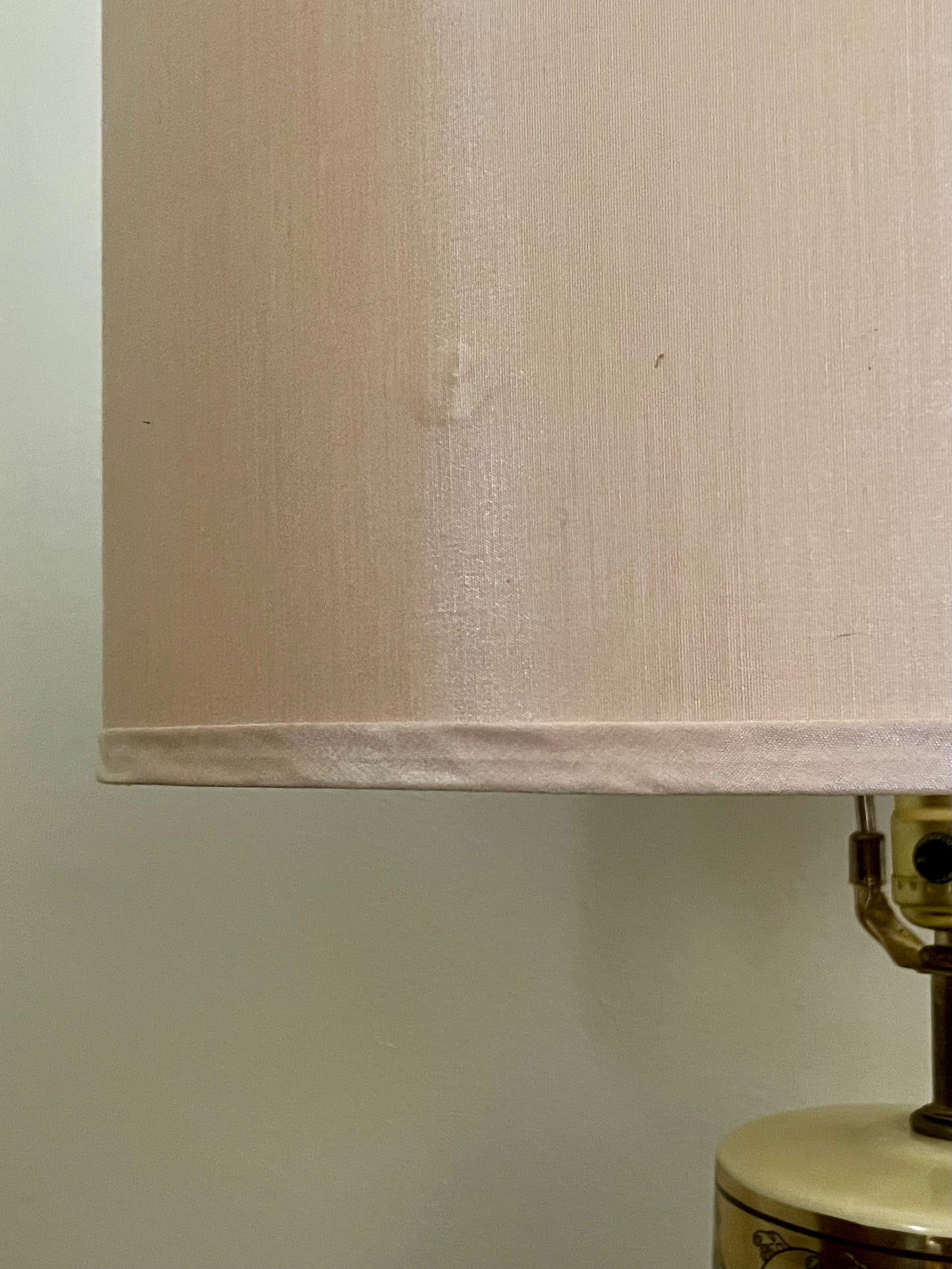 A close-up of a light beige silk fabric lampshade with a small visible dent, attached to a brass and cream-colored lamp base against a plain off-white wall.