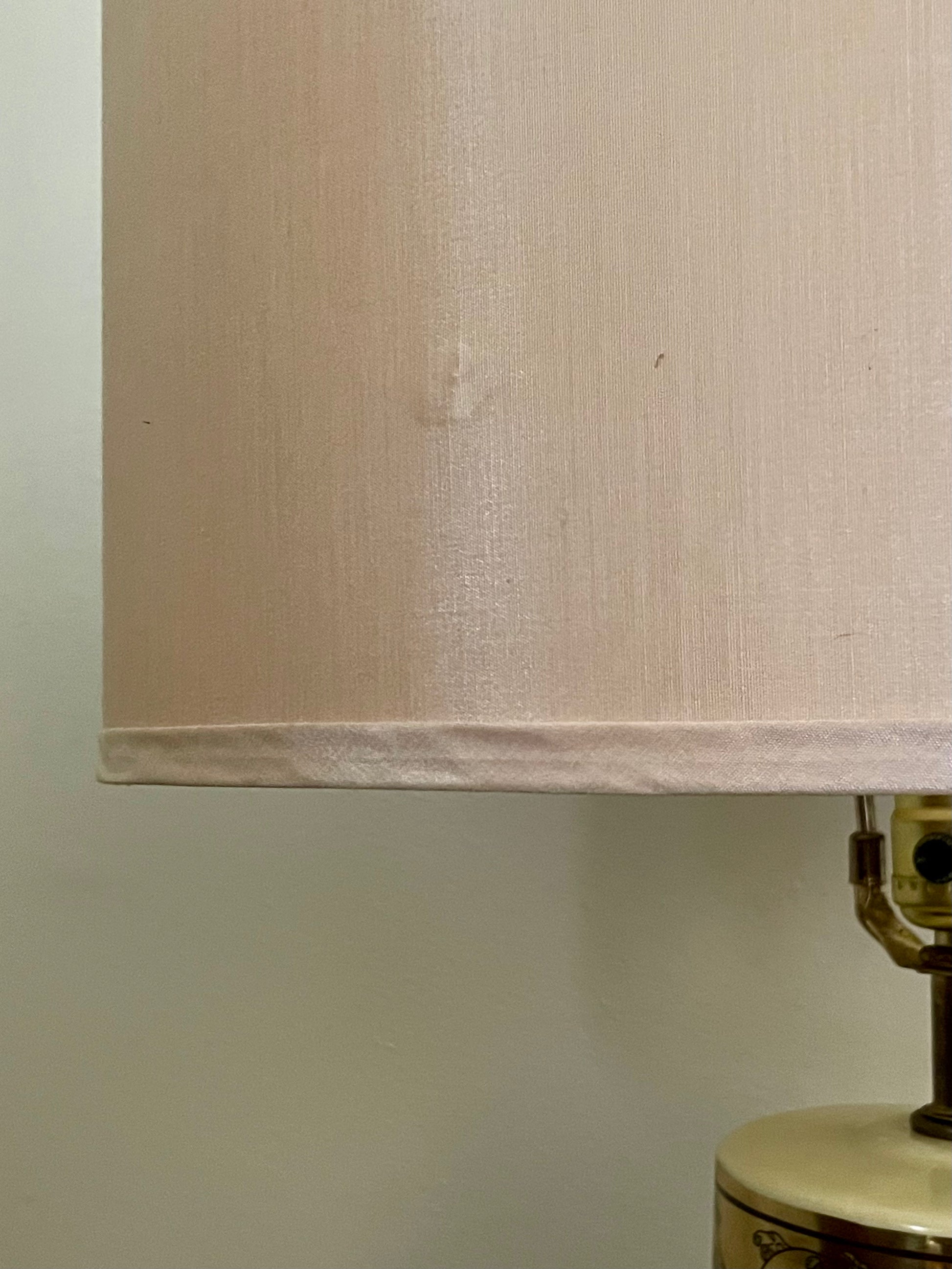 A close-up of a light beige silk fabric lampshade with a small visible dent, attached to a brass and cream-colored lamp base against a plain off-white wall.