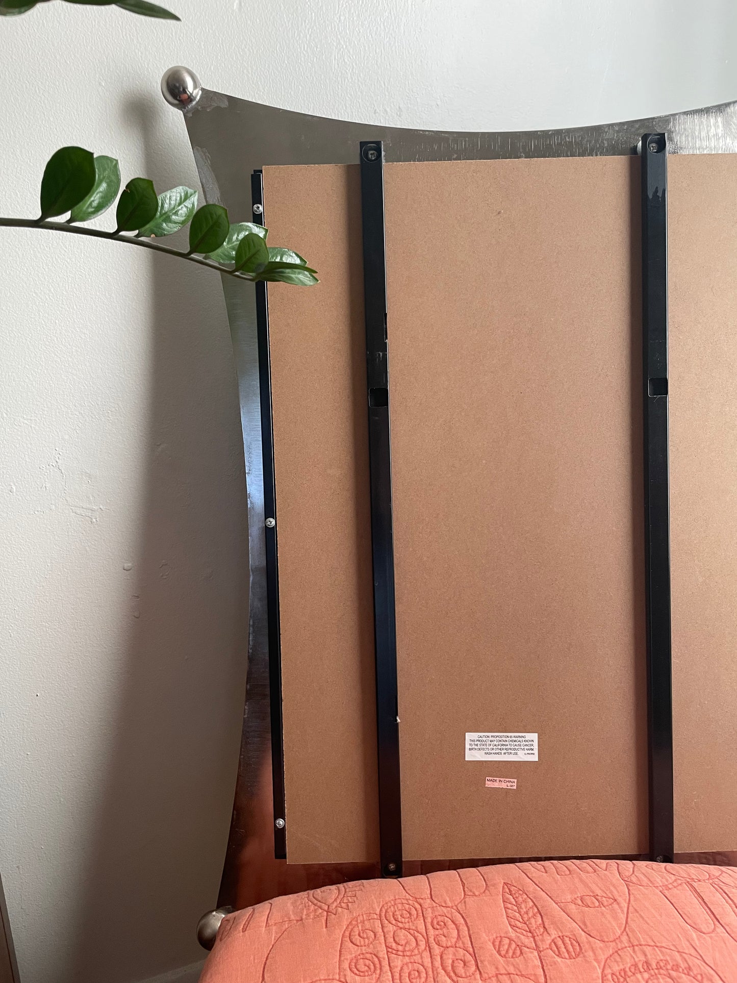 The back view of a silver framed mirror, showing its brown cardboard backing panels and black metal support brackets, set against a white wall with a plant leaf on the left.