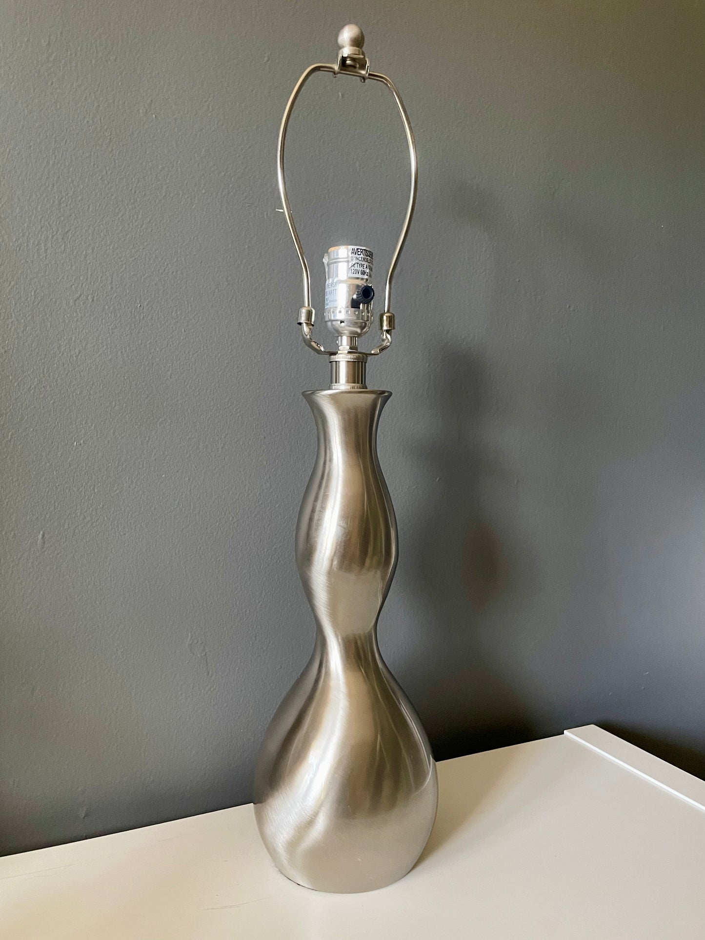 A table lamp with a sculptural, hourglass-shaped metallic silver base, set on a white surface against a dark gray wall.