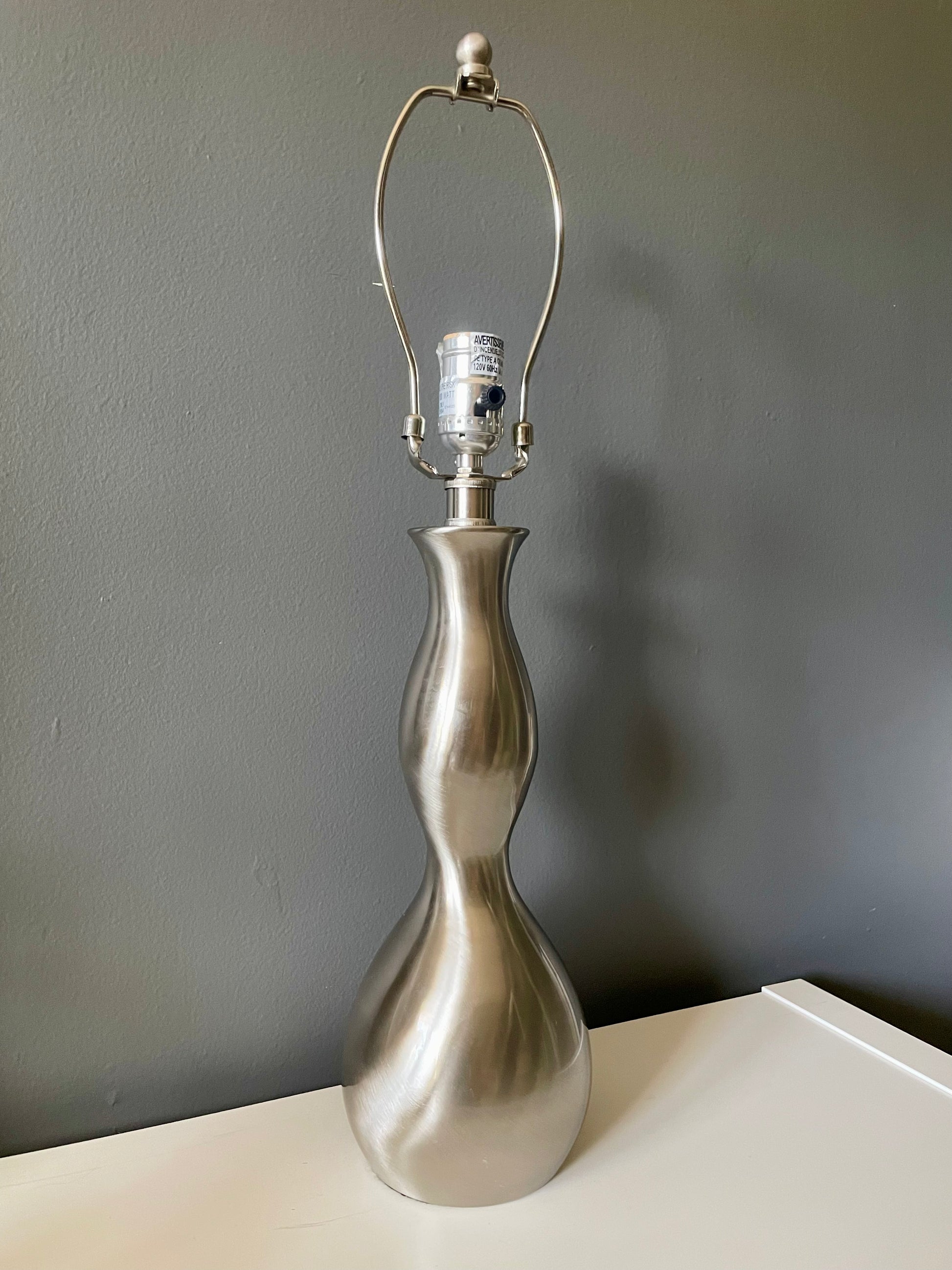A table lamp with a sculptural, hourglass-shaped metallic silver base, set on a white surface against a dark gray wall.