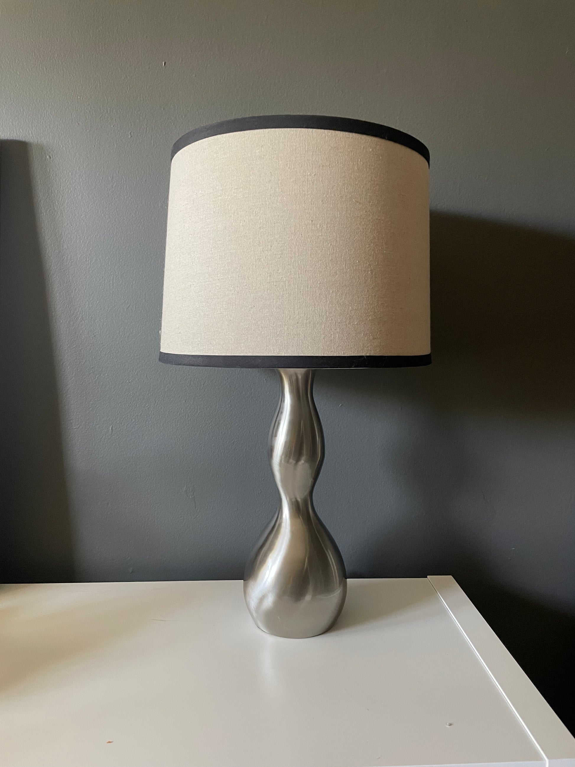 A table lamp with a textured beige fabric drum shade trimmed in black, mounted on a sculptural, hourglass-shaped metallic silver base, set on a white surface against a dark gray wall.