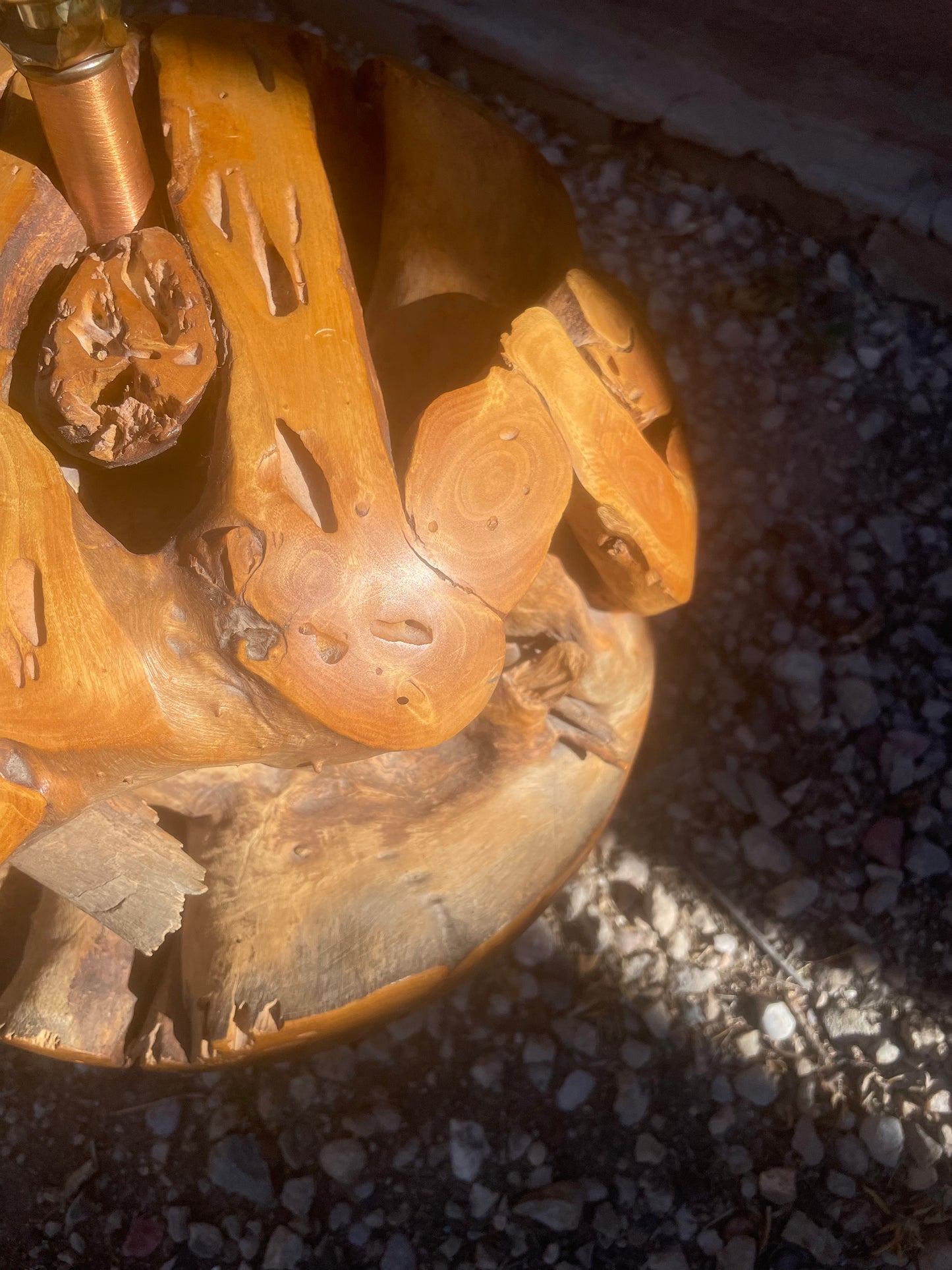 A close-up of a sculptural polished wood lamp showing the natural variation in grain and organic shape of a root ball, shown outdoors on gravel under direct sunlight.