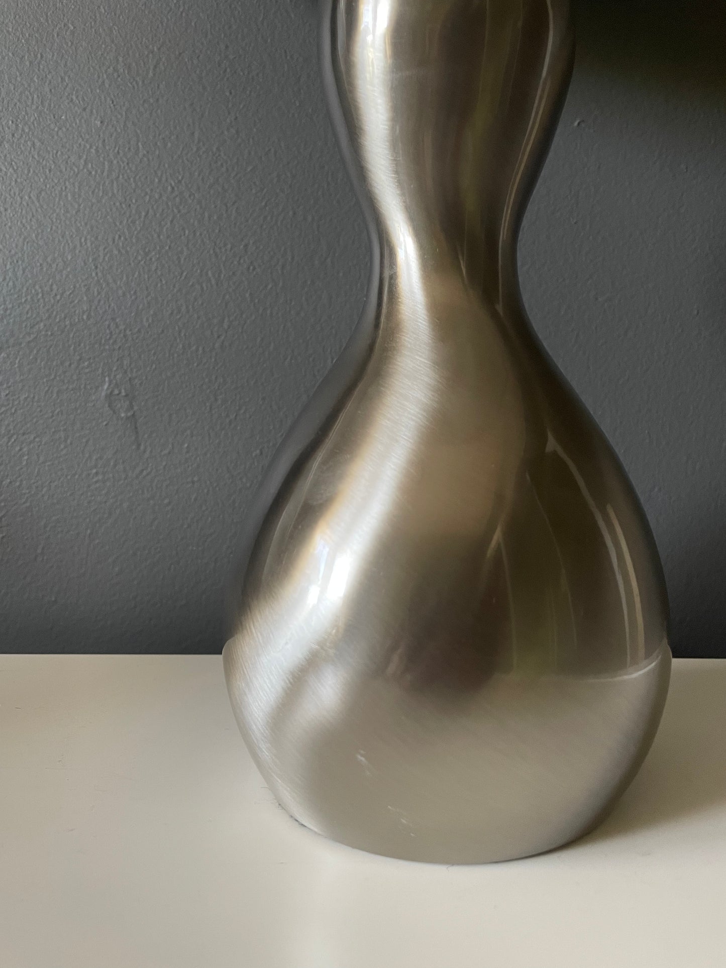 Closeup of a table lamp with a sculptural, hourglass-shaped metallic silver base, set on a white surface against a dark gray wall.