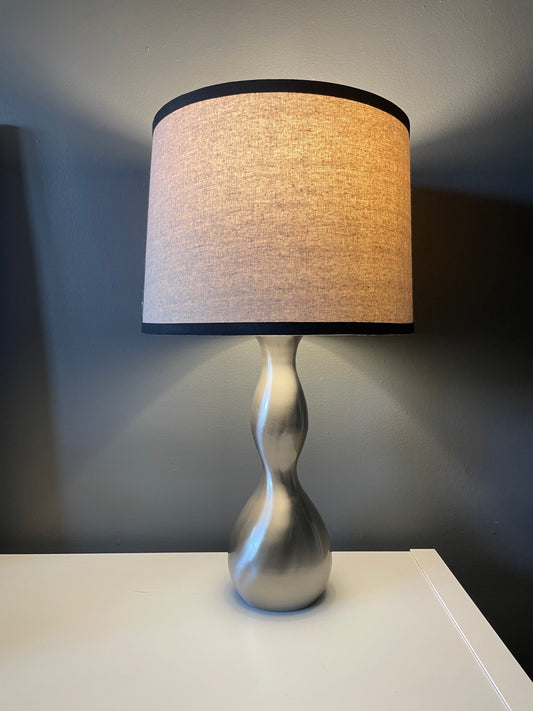 A lit table lamp with a textured beige fabric drum shade trimmed in black, mounted on a sculptural, hourglass-shaped metallic silver base, set on a white surface against a dark gray wall.