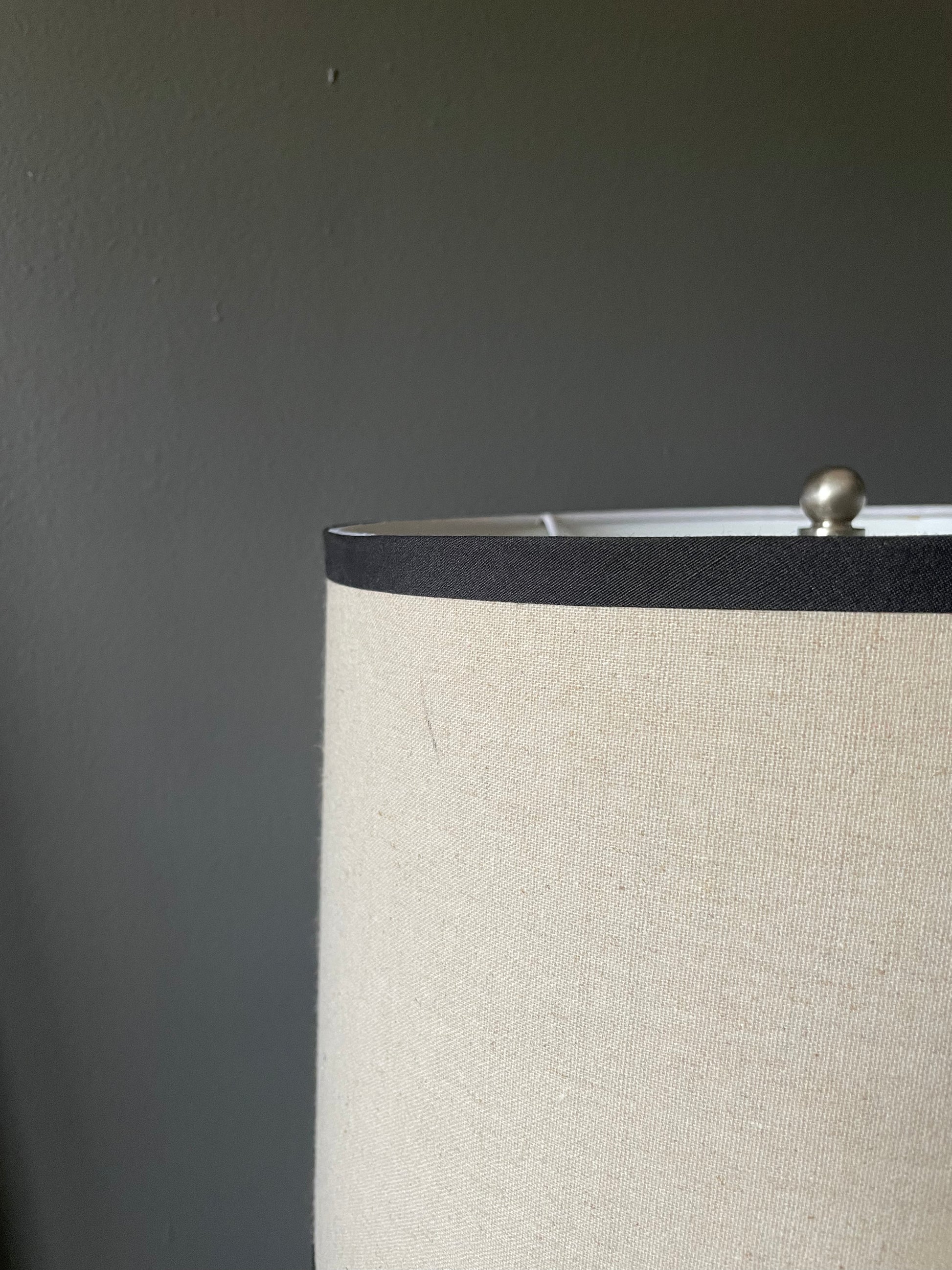 Close-up of a lamp shade with a textured beige fabric exterior and a black band near the top, set against a dark gray wall.
