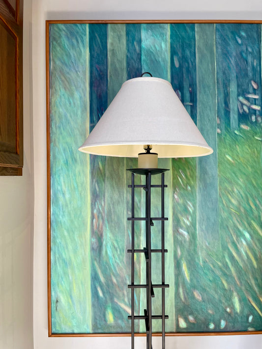 A floor lamp with a large white conical shade and a black iron frame, positioned in front of a large framed painting featuring vertical strokes of teal, green, and blue.