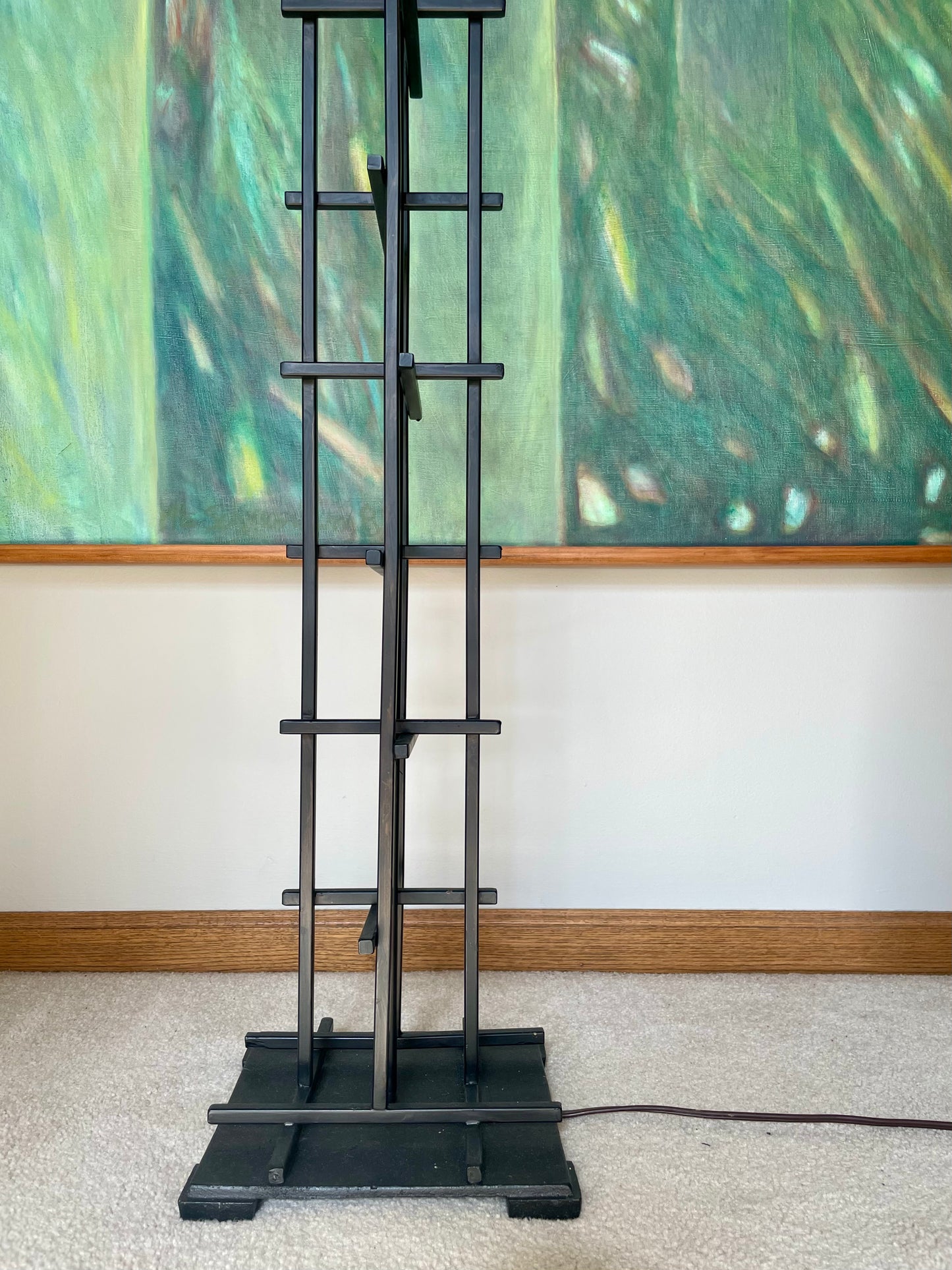 A tall black metal floor lamp with a vertical frame, horizontal support bars, and a square base, standing on a beige carpet in front of a white wall and a large green abstract painting.