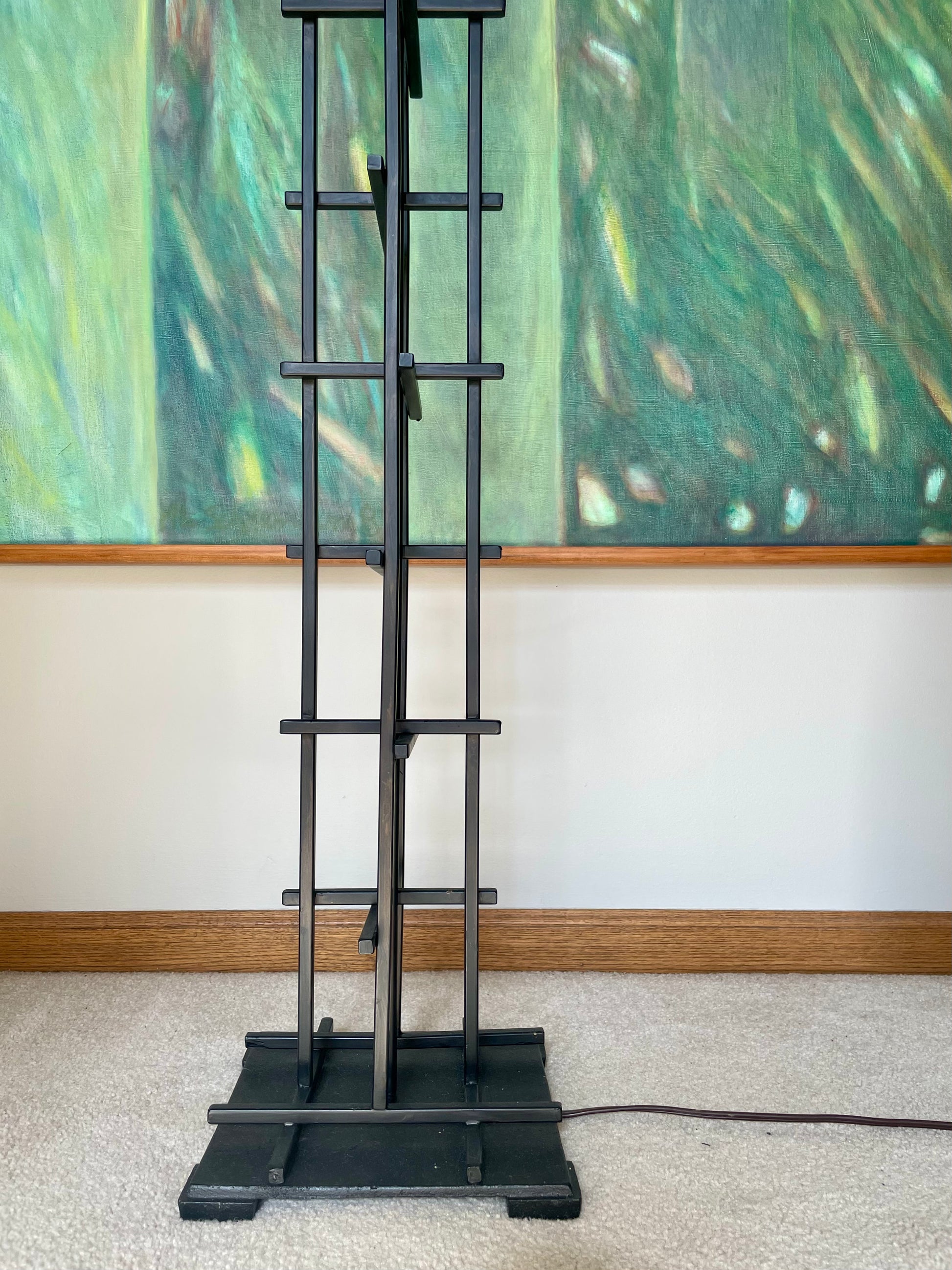A tall black metal floor lamp with a vertical frame, horizontal support bars, and a square base, standing on a beige carpet in front of a white wall and a large green abstract painting.