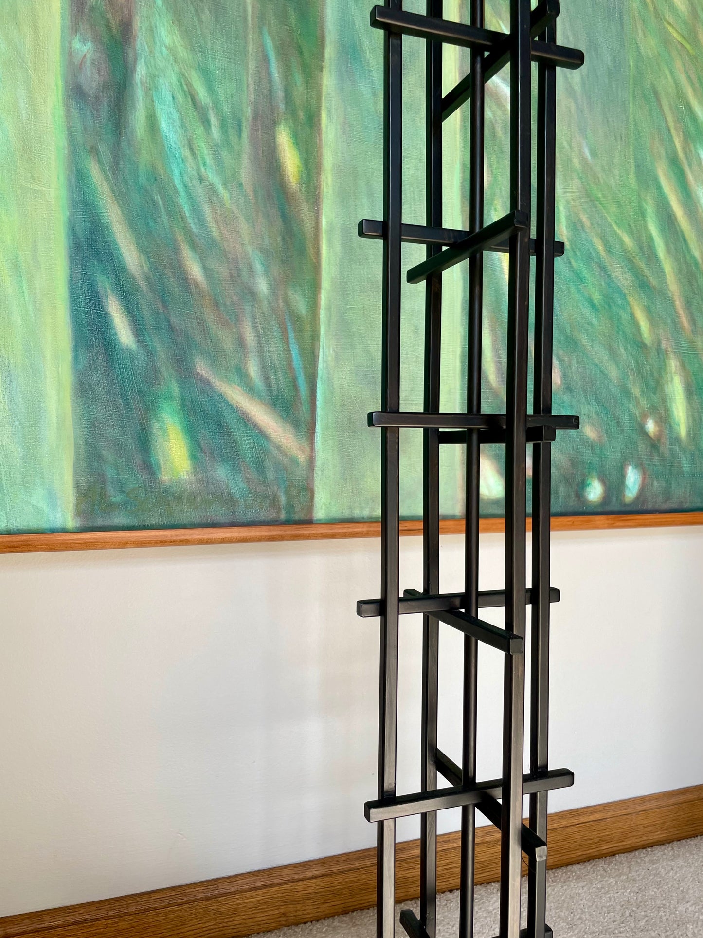 A tall black iron grid-style shelving unit with multiple tiers, positioned in front of a large green abstract painting on a white wall.