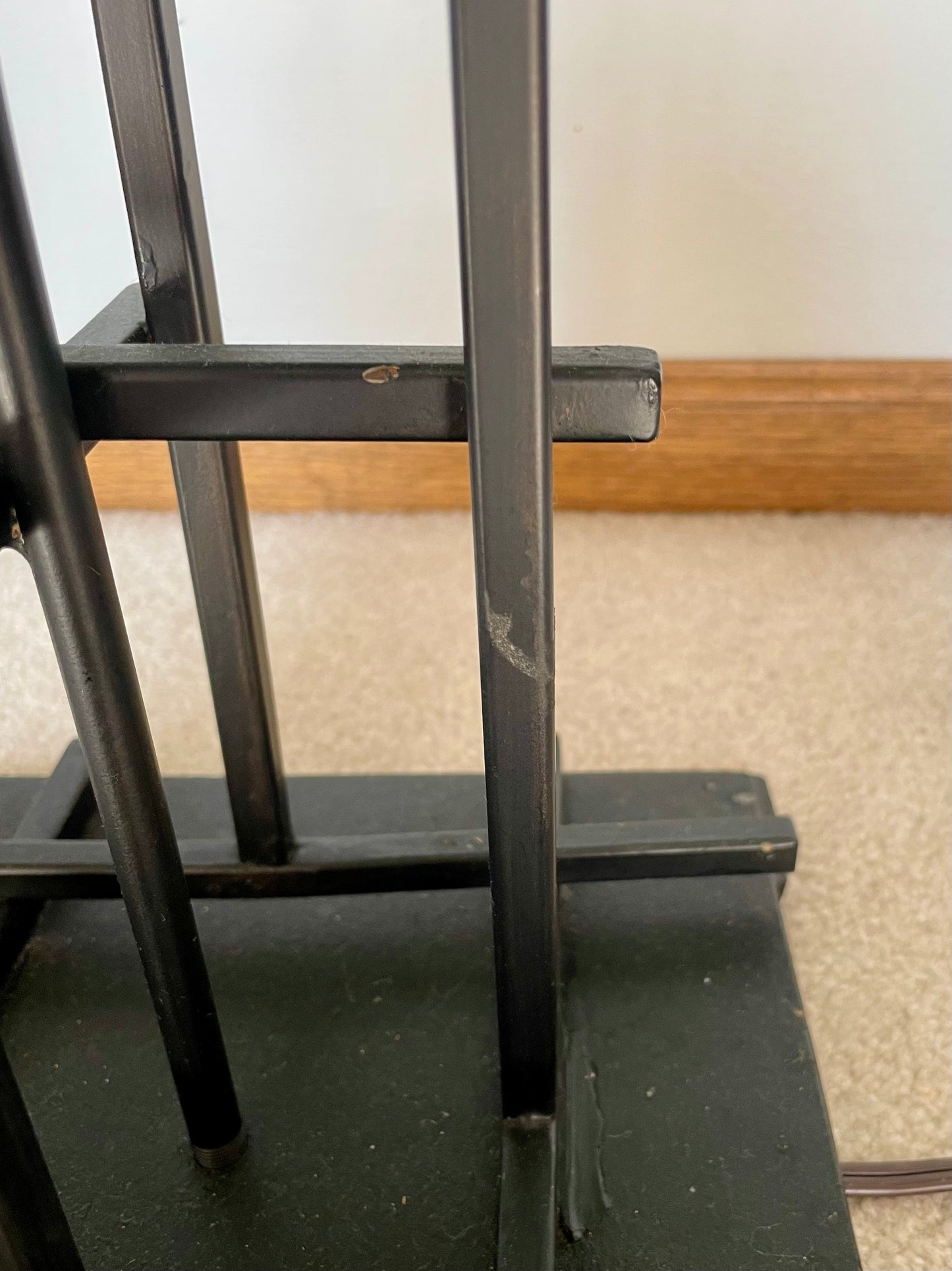 Close-up of a black metal stand or frame structure with vertical bars and horizontal supports, photographed indoors on a carpeted floor against a light wall.