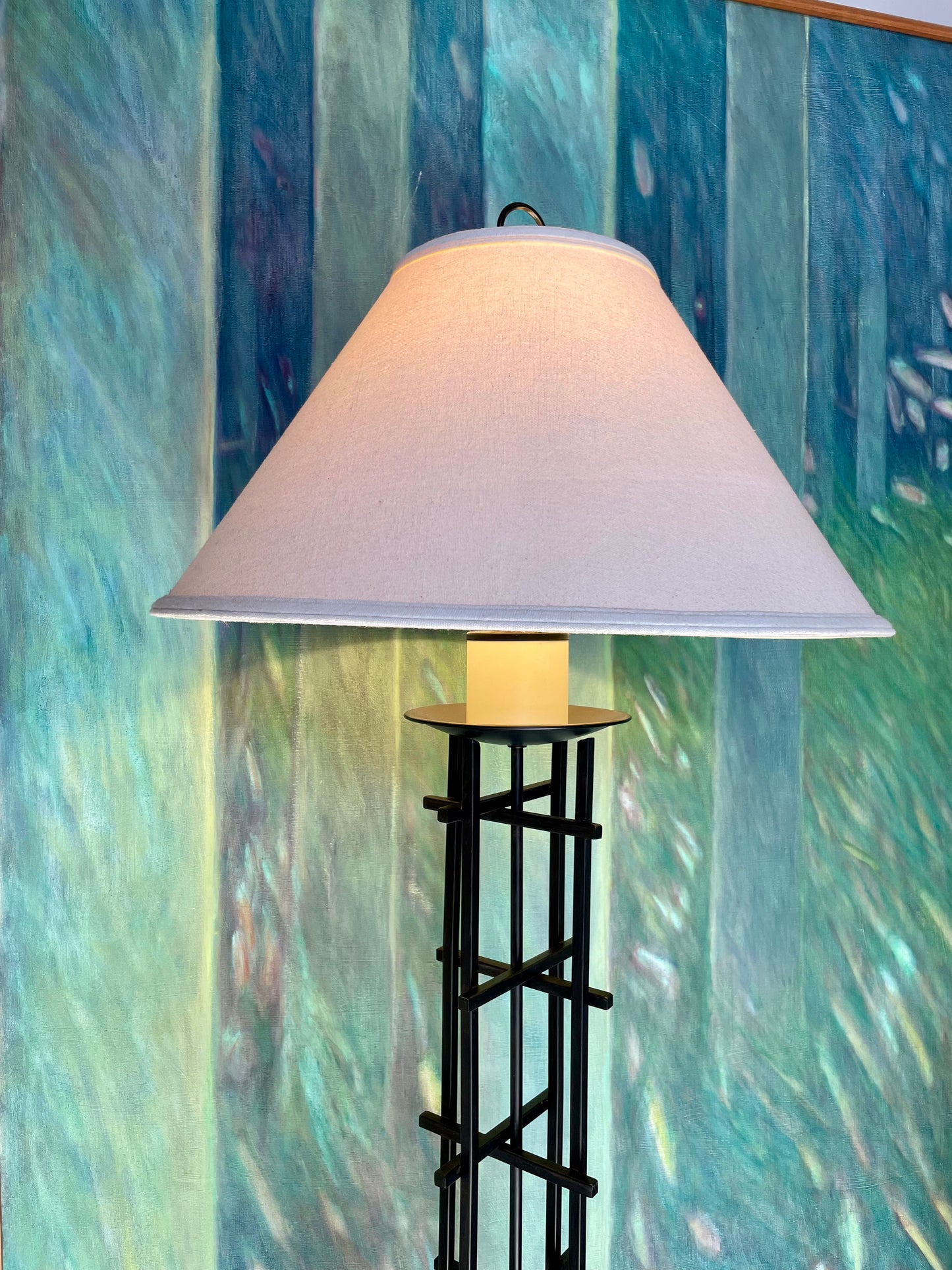 A floor lamp with a large tapered off-white linen shade, a lit bulb, and a black metal frame, set against a textured blue and green abstract wall backdrop.