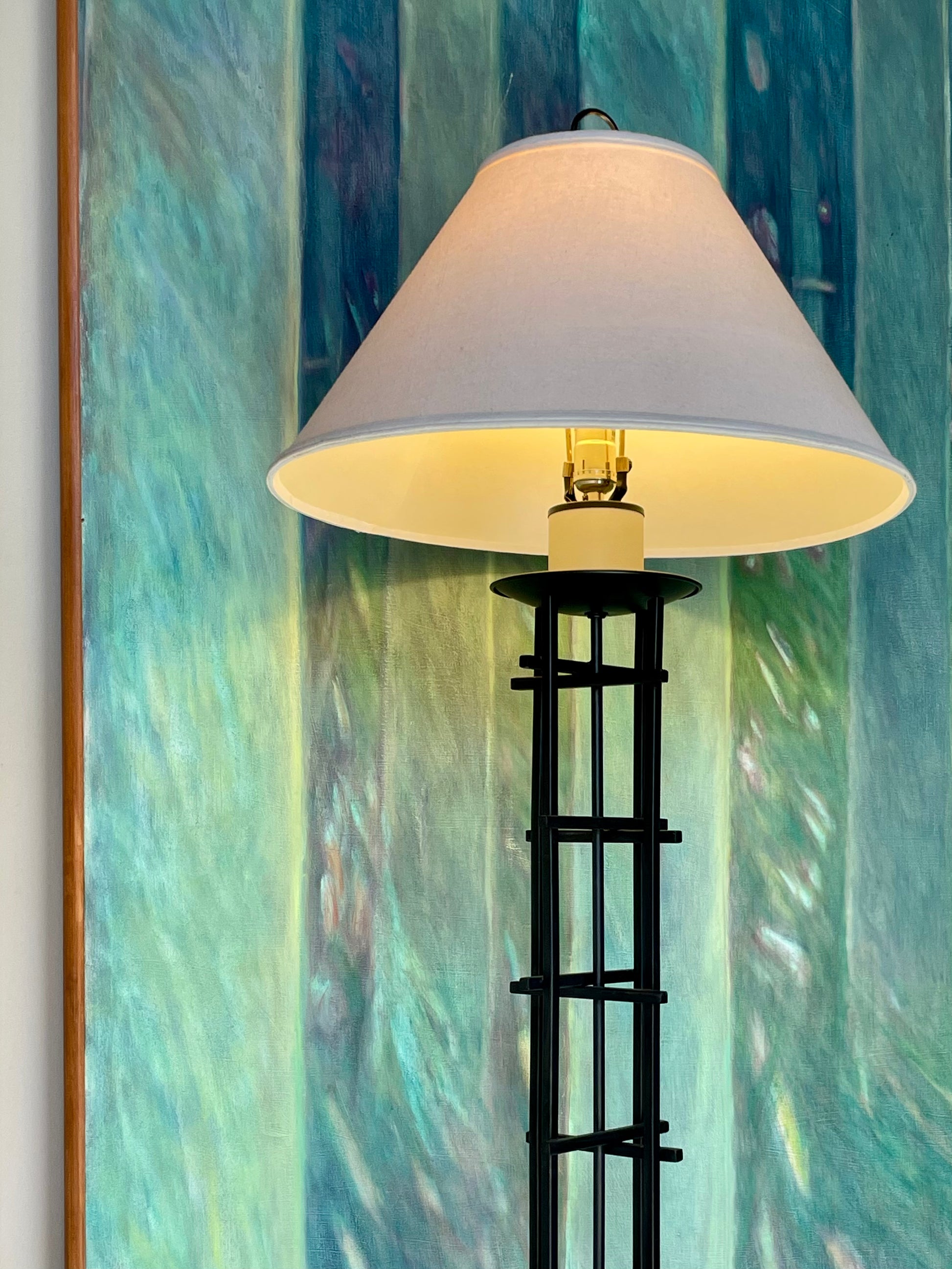 A floor lamp with a large white conical shade and a black iron frame, illuminated against a textured blue and green abstract wall backdrop.