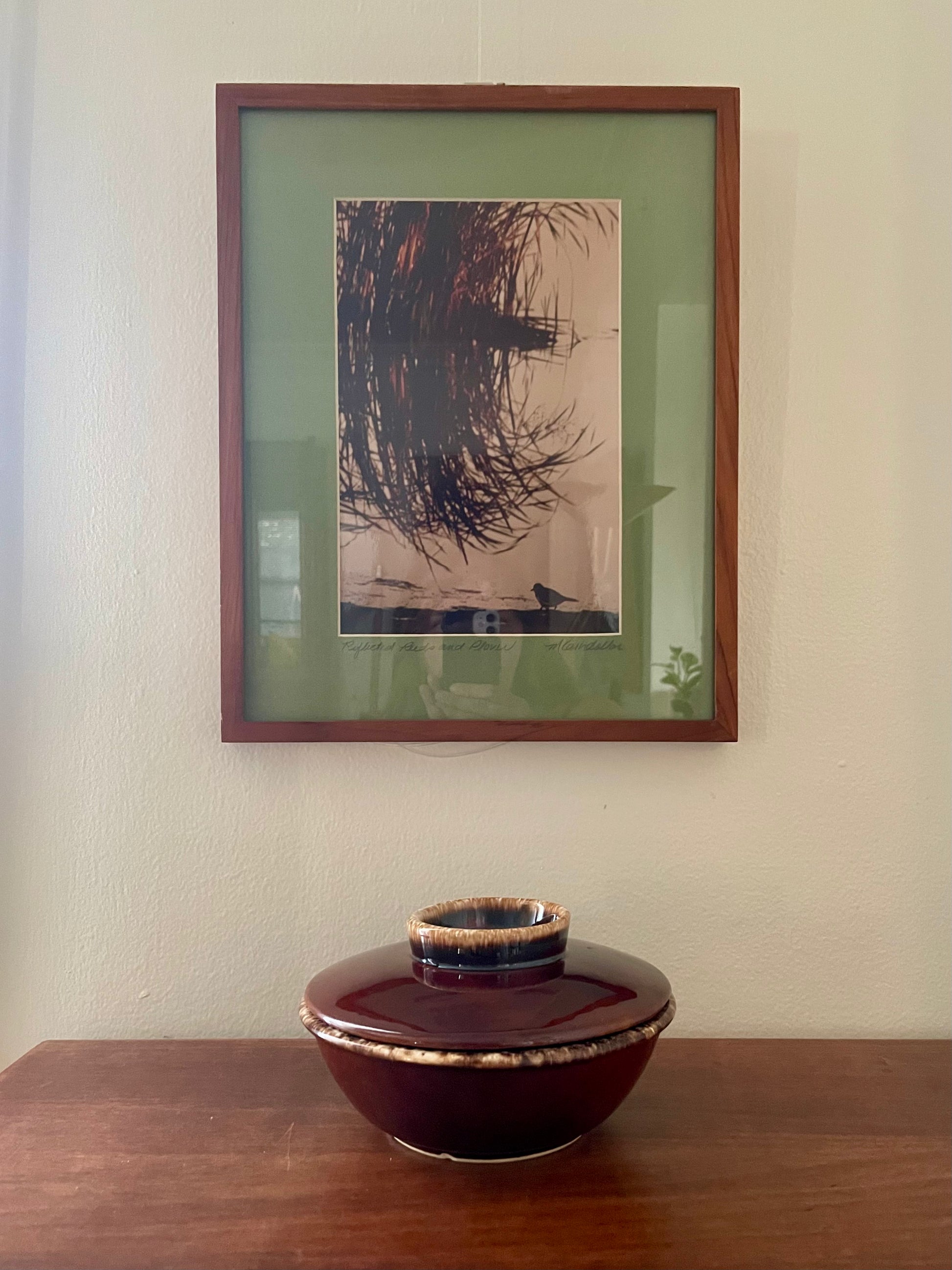 A framed photograph of curved reeds reflecting in the water with a plover nearby with a green mat and dark wood frame hangs on a beige wall above a wooden table with a dark glazed ceramic lidded container.
