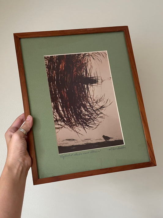 A hand holds a framed art print featuring a silhouette of reeds and a plover on a reflective water surface, set against a muted green mat and a warm brown wooden frame.