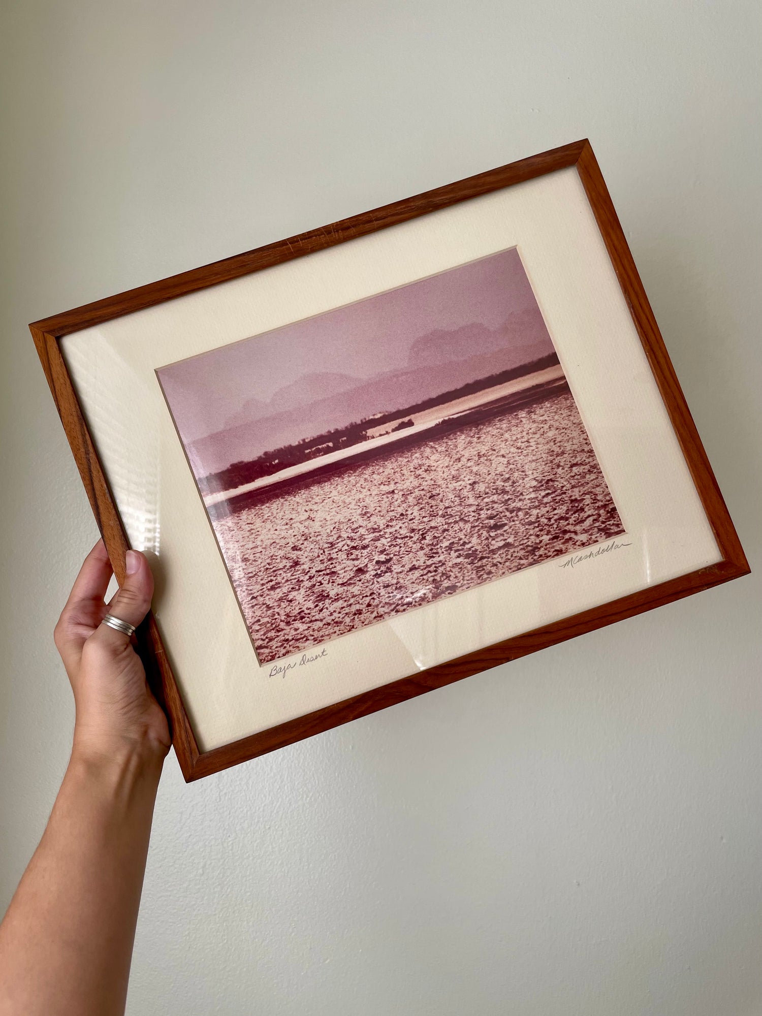 A framed art print featuring a pink-toned landscape photograph, held by a hand against a plain white wall. The print is matted with cream and enclosed in a medium brown wooden frame.