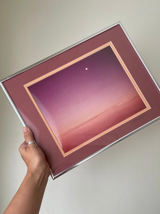A hand holding a square framed photograph with a silver frame, a mauve mat, and a central landscape print featuring a sunset gradient from purple to pink and orange with a moon in the sky.