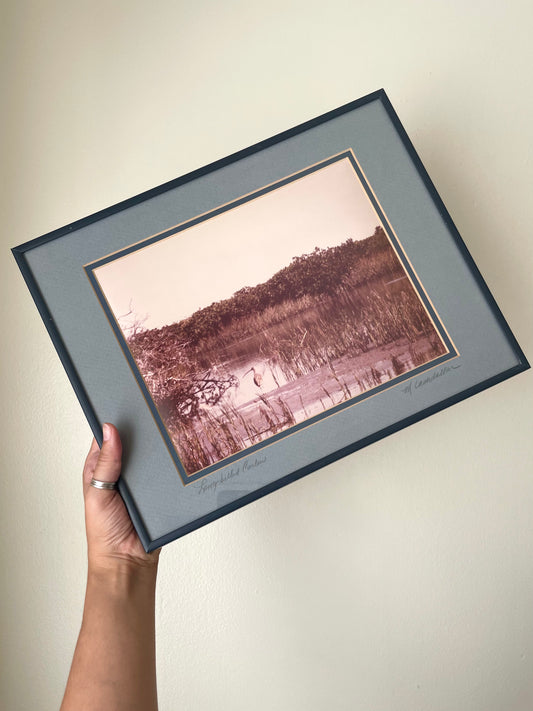 A hand holds a framed art print against a plain beige wall. The print features a sepia-toned landscape photograph with a long-billed curlew, surrounded by a wide blue-gray mat, narrow denim blue border, and a dark navy blue frame.