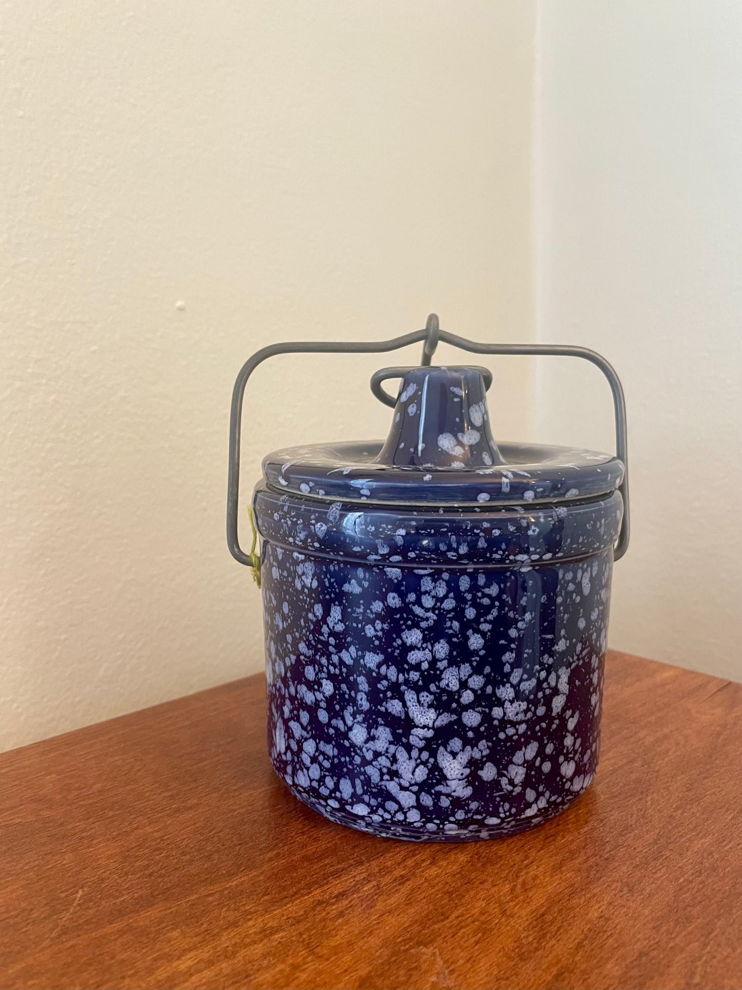 A round vintage stoneware lidded container with a dark metal wire handle, featuring a blue and white speckled pattern, sitting on a wooden table against a beige wall.