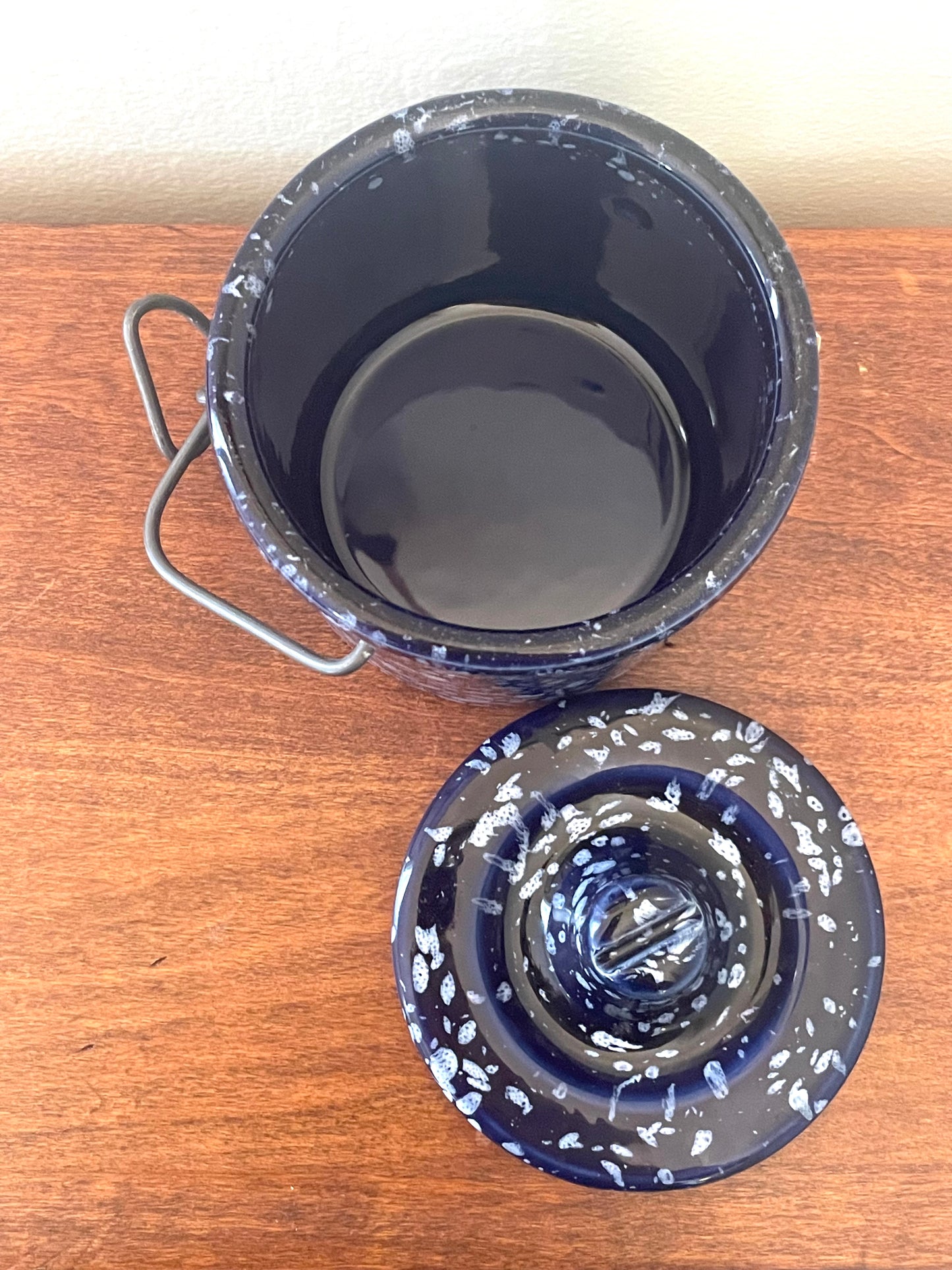 A dark blue stoneware lidded container with white speckling, shown open on a wooden tabletop with its metal handle visible and the matching lid placed in front.