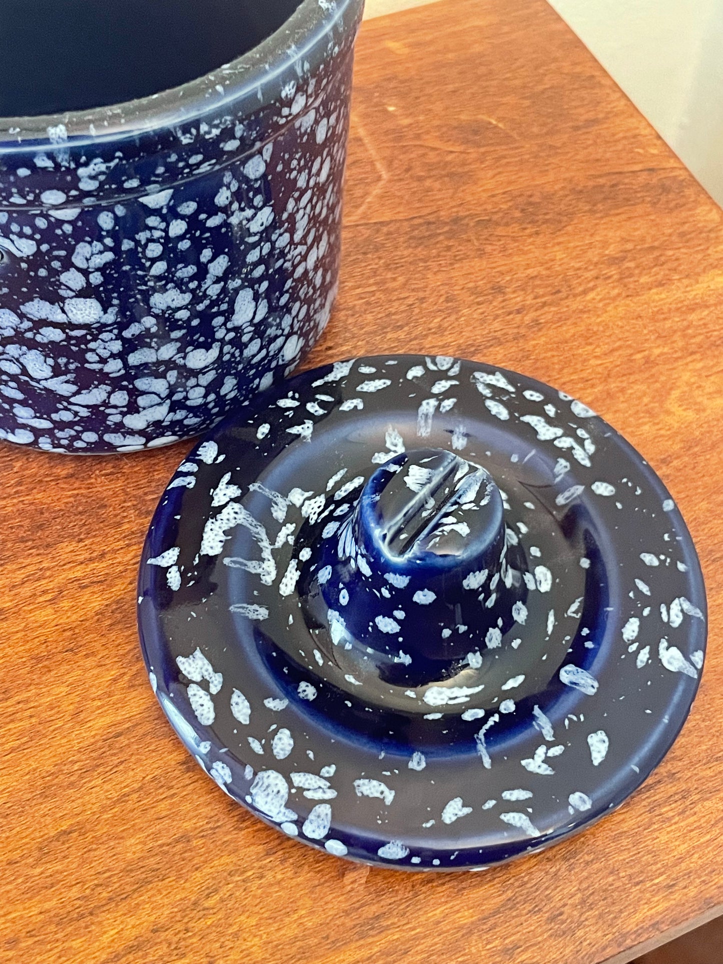 A dark blue and white speckled ceramic lidded container shown on a wooden tabletop, with the lid placed in front and part of the container visible behind it.