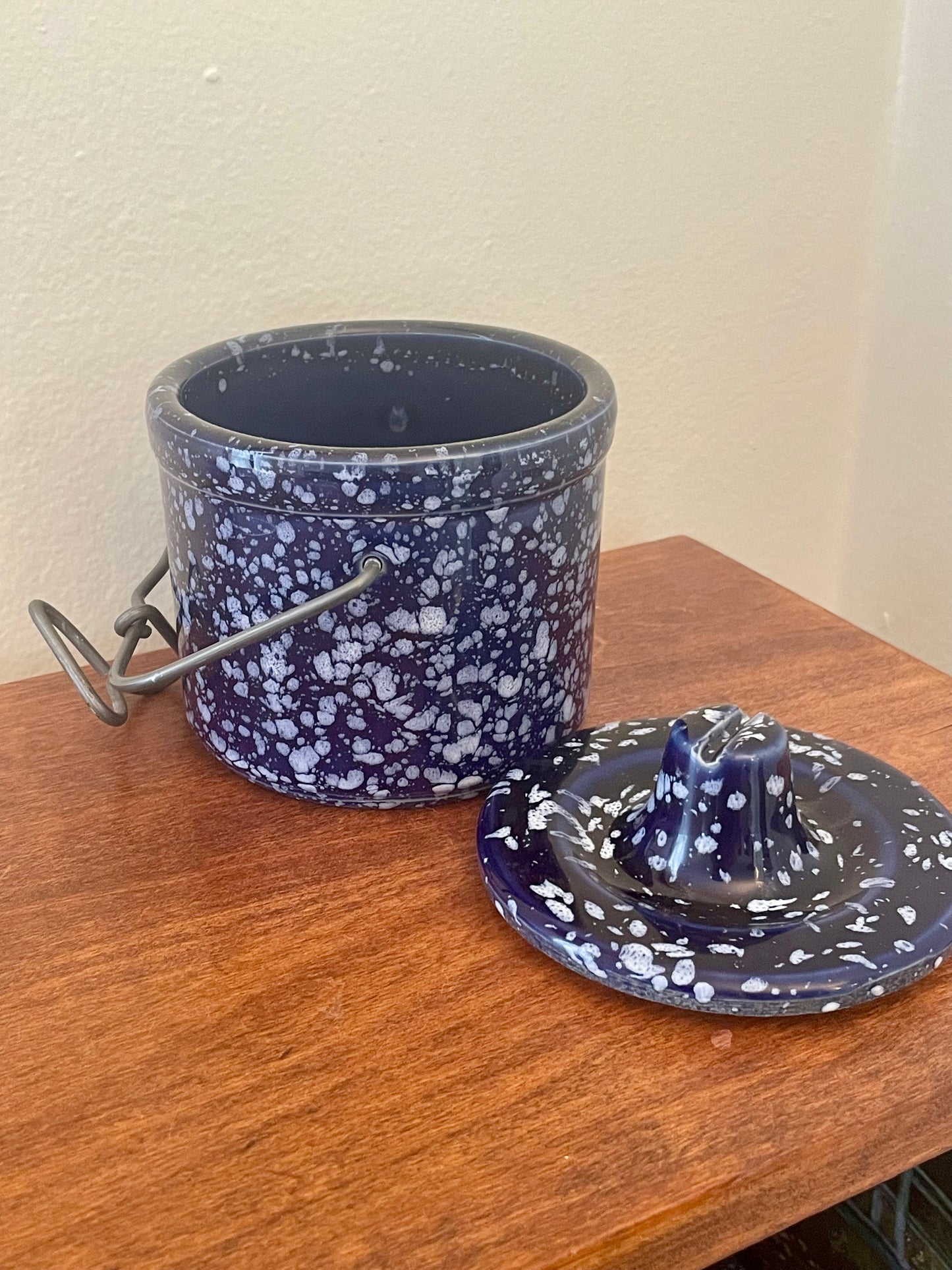 A dark blue and white speckled stoneware storage container with a metal handle, shown next to its matching lid on a wooden surface against a beige wall.