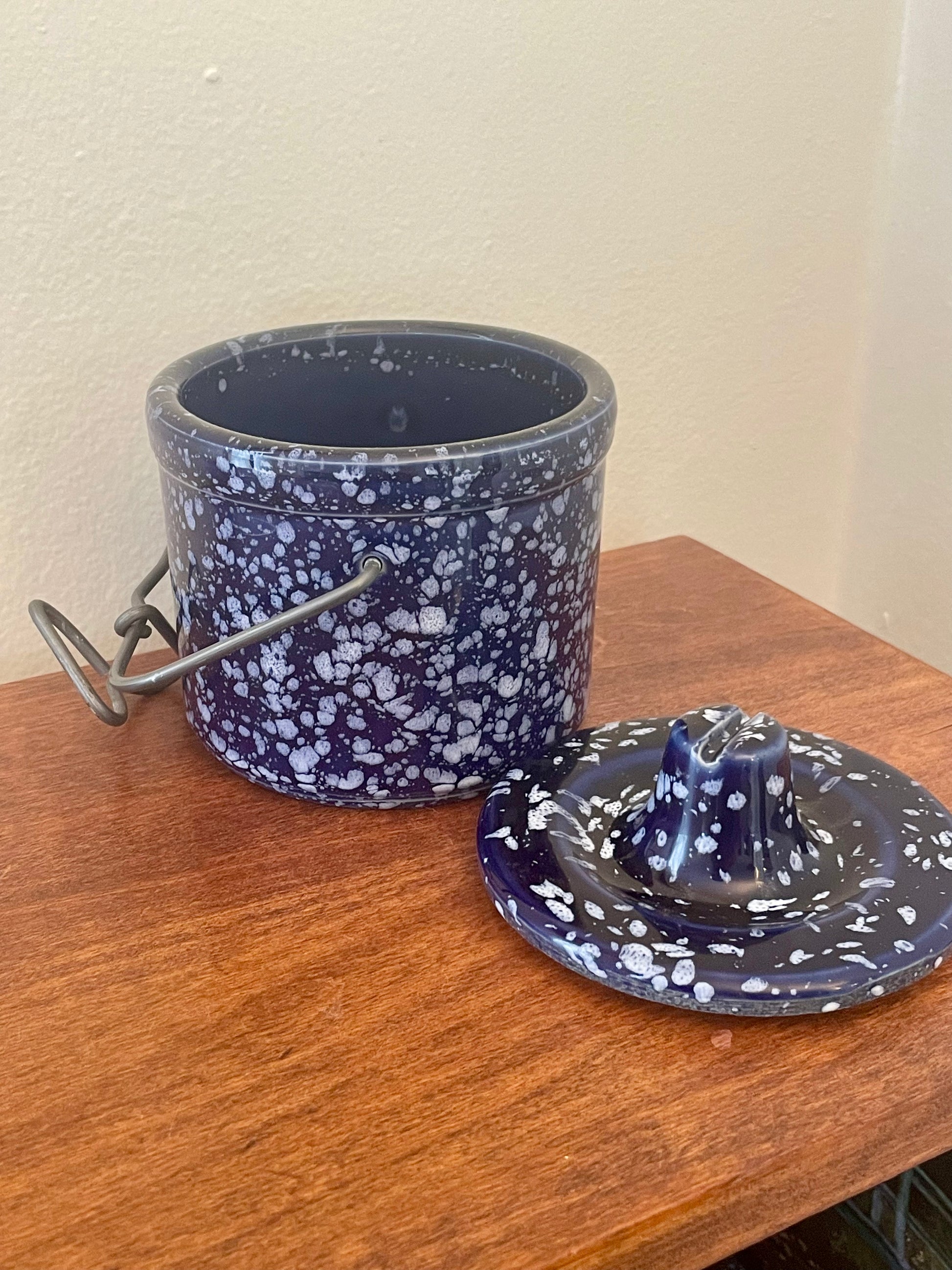 A dark blue and white speckled stoneware storage container with a metal handle, shown next to its matching lid on a wooden surface against a beige wall.