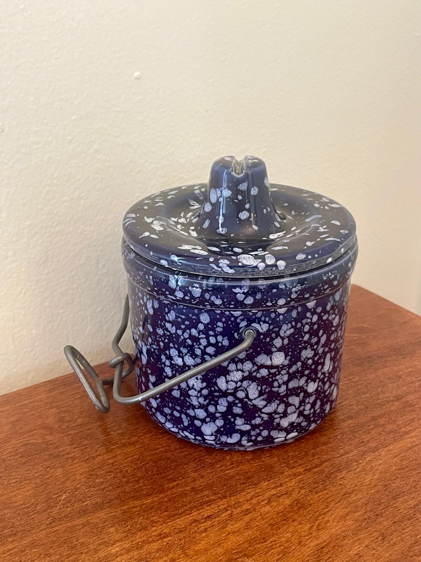 A vintage ceramic lidded container with a dark blue and white speckled glaze, featuring a metal wire handle, resting on a wooden surface against a plain wall.