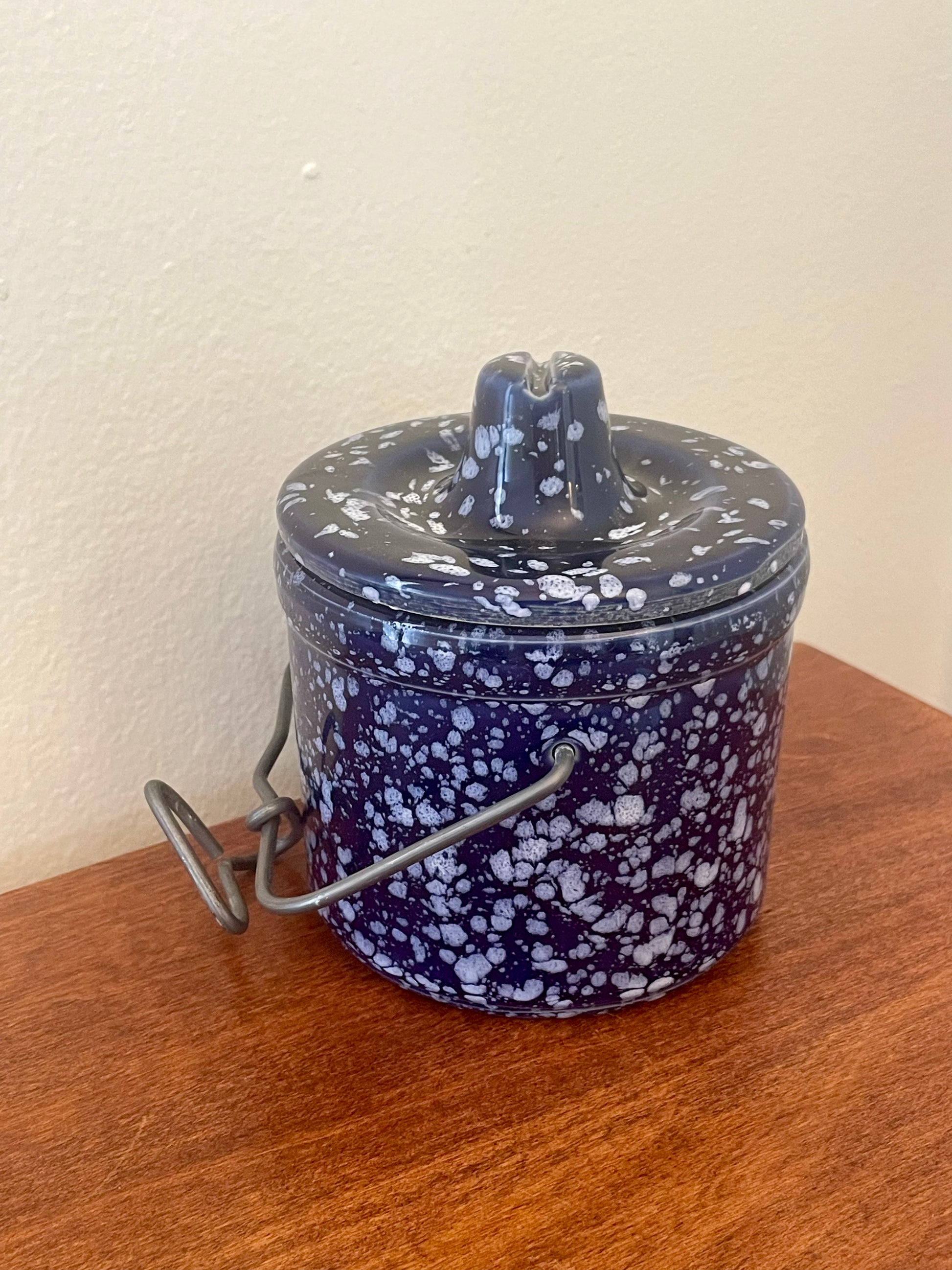 A vintage ceramic lidded container with a dark blue and white speckled glaze, featuring a metal wire handle, resting on a wooden surface against a plain wall.