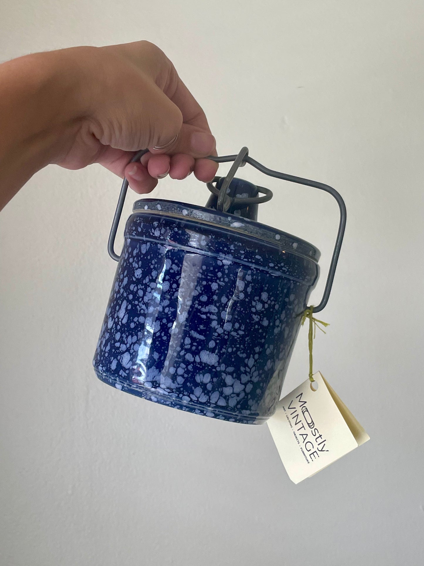 A hand holding a small blue stoneware canister with white speckles, a dark metal handle, and a hanging paper tag that reads 'Mostly Vintage'.
