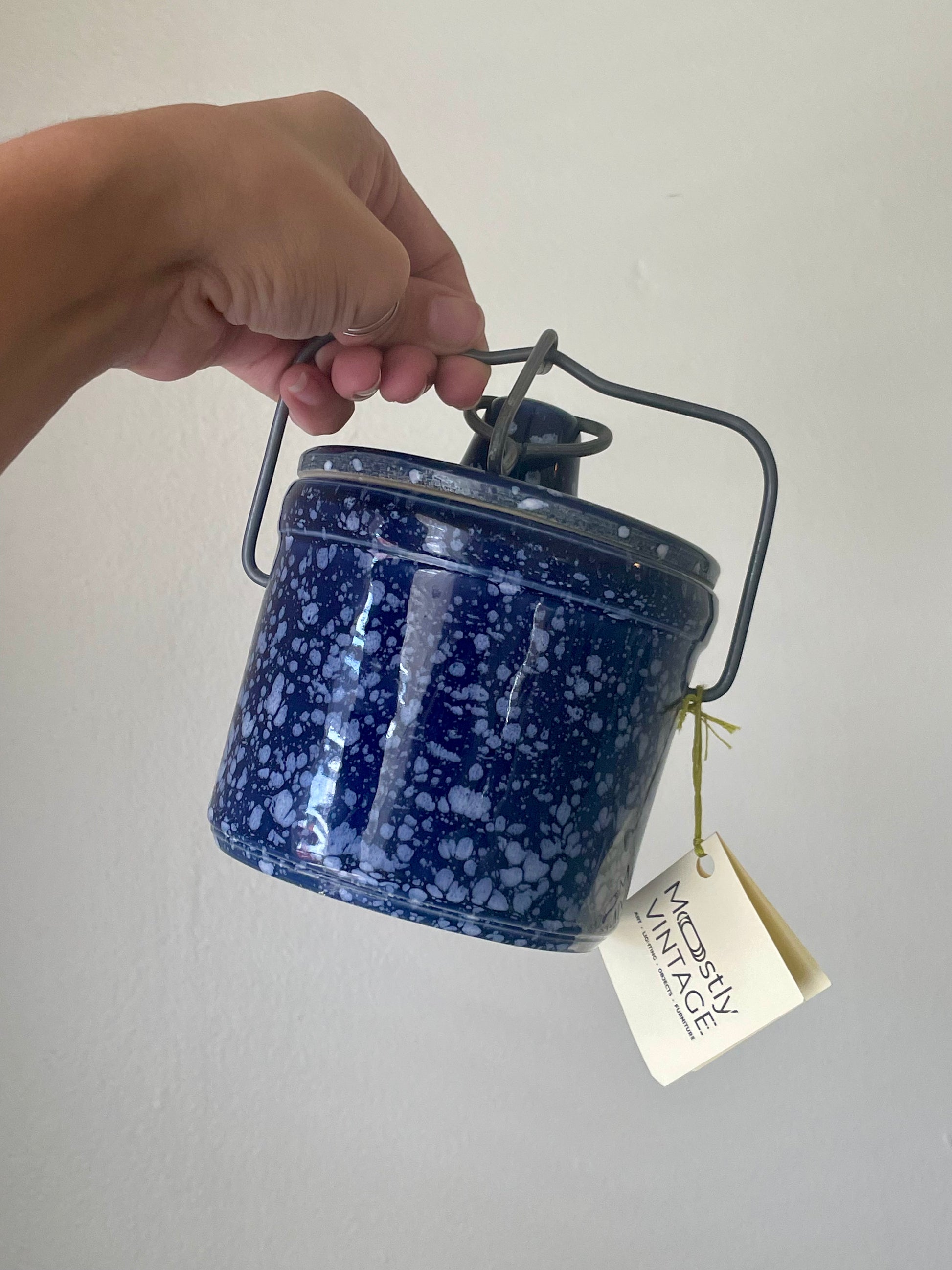 A hand holding a small blue stoneware canister with white speckles, a dark metal handle, and a hanging paper tag that reads 'Mostly Vintage'.