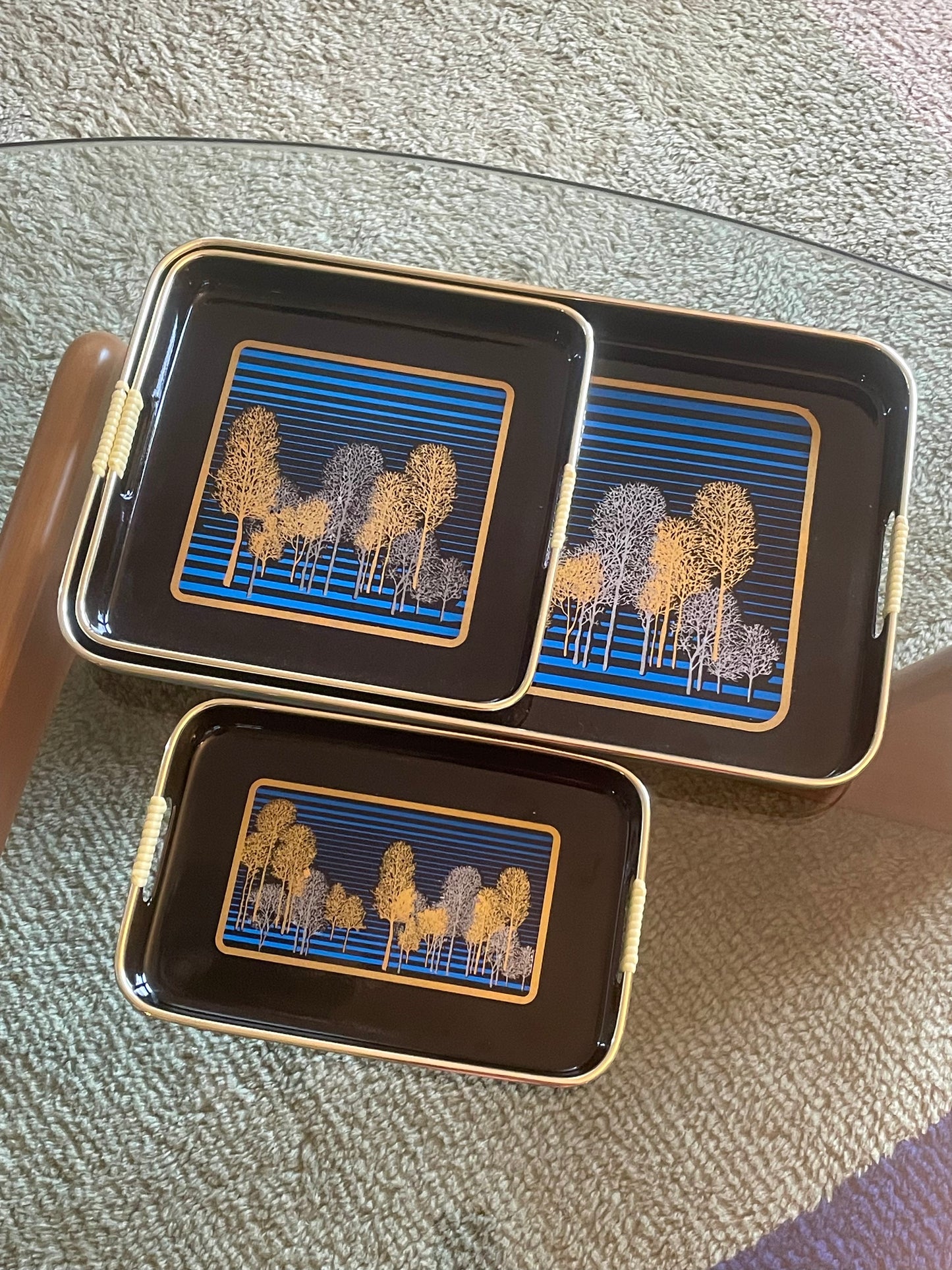 Three rectangular black serving trays with gold-toned edges and handles, featuring a blue striped and gold and silver tree pattern, arranged on a glass table over a textured rug.