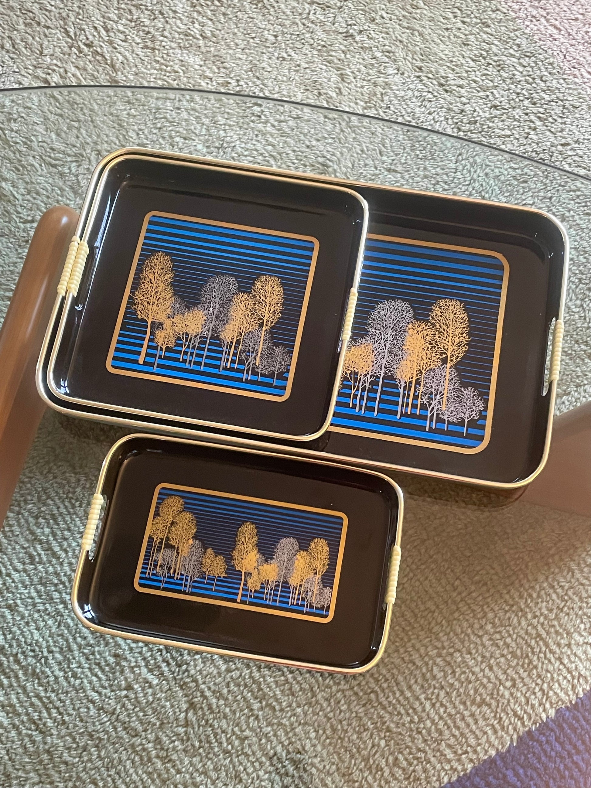 Three rectangular black serving trays with gold-toned edges and handles, featuring a blue striped and gold and silver tree pattern, arranged on a glass table over a textured rug.